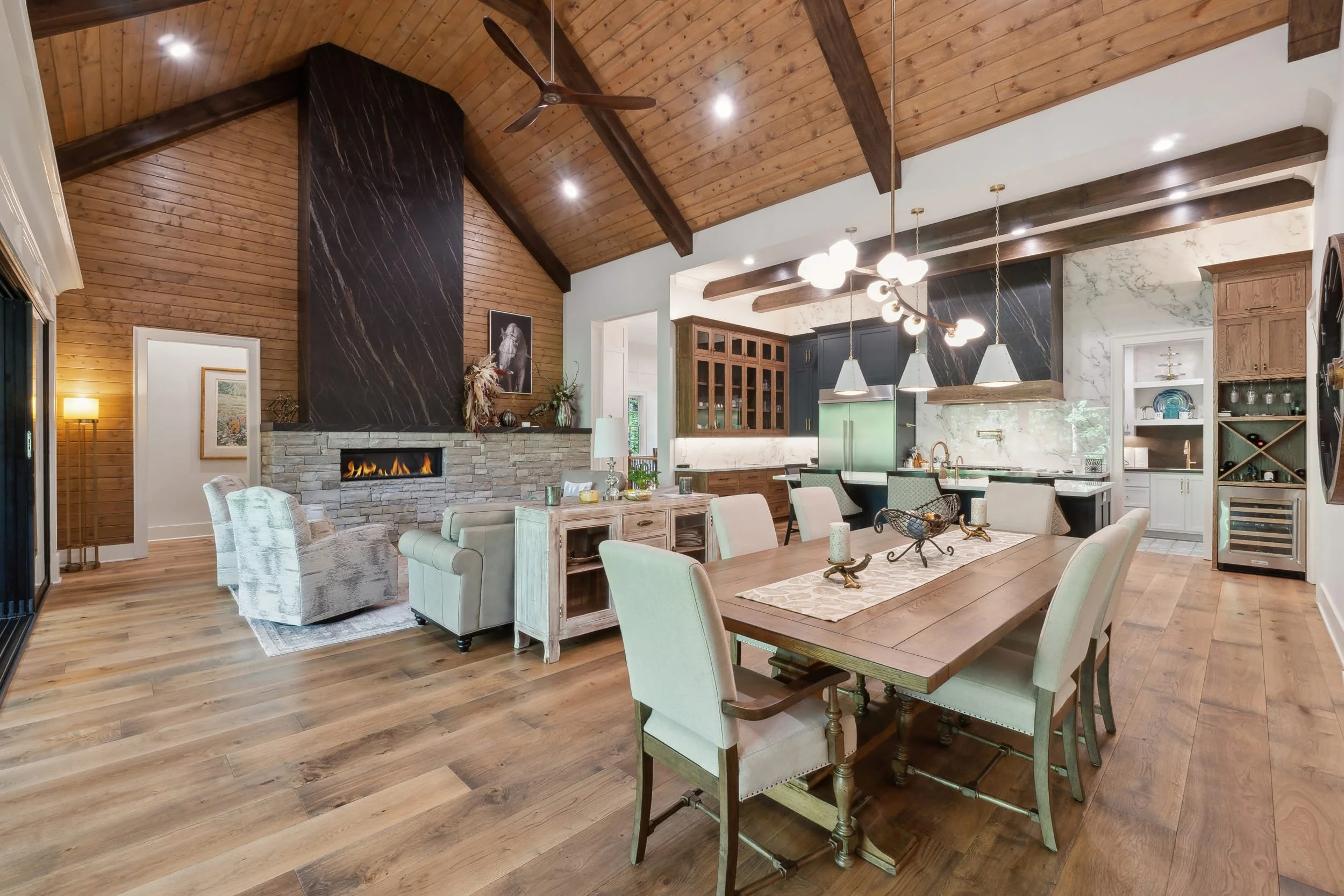 Interior of a spacious living and kitchen area with a vaulted wood ceiling, wooden beams, a fireplace, dining table with chairs, modern lighting, and kitchen cabinets.