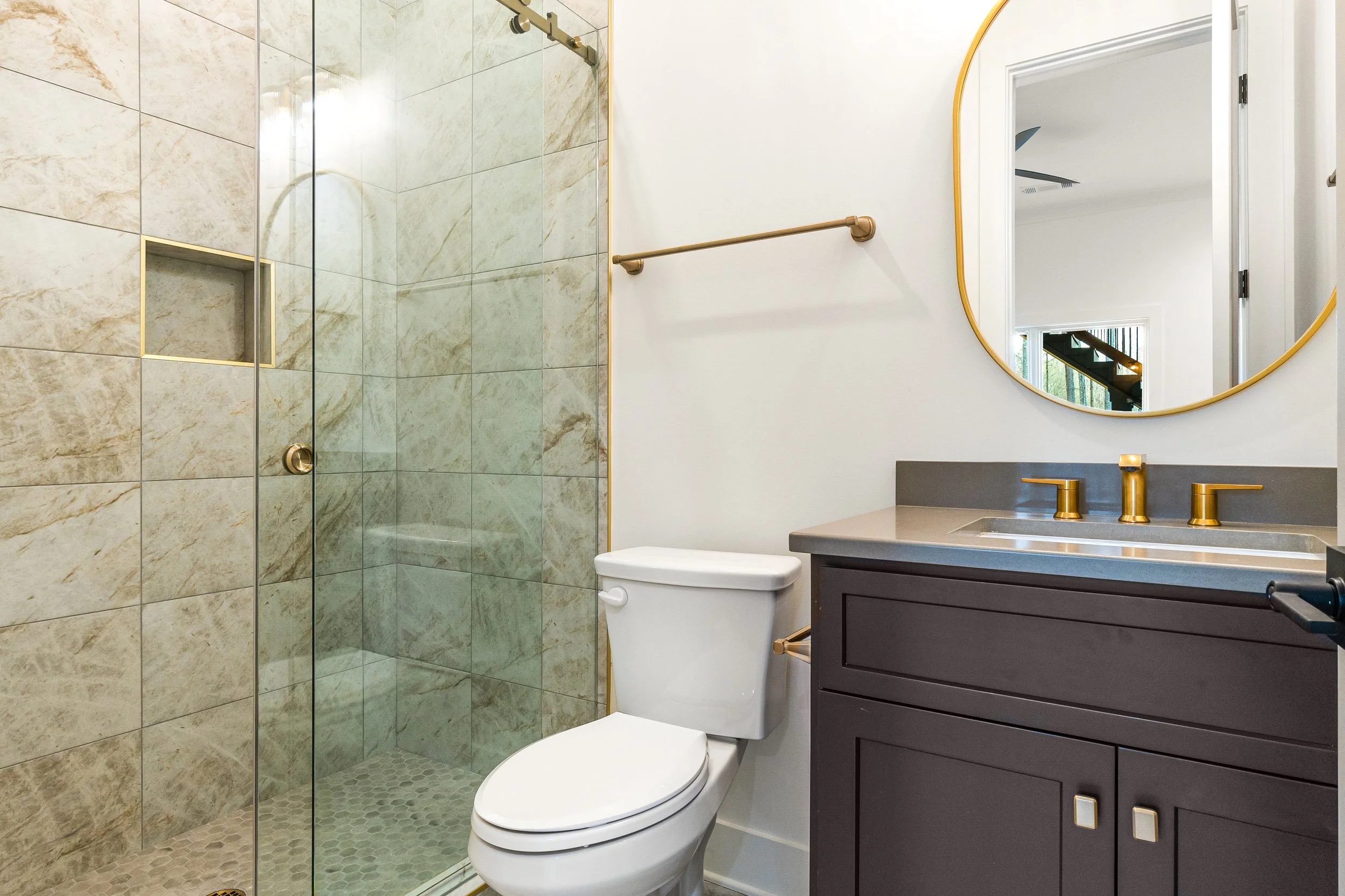 Modern bathroom with a glass shower enclosure, a white toilet, and a gray vanity with gold fixtures, a large round mirror, and a window showing a staircase outside.