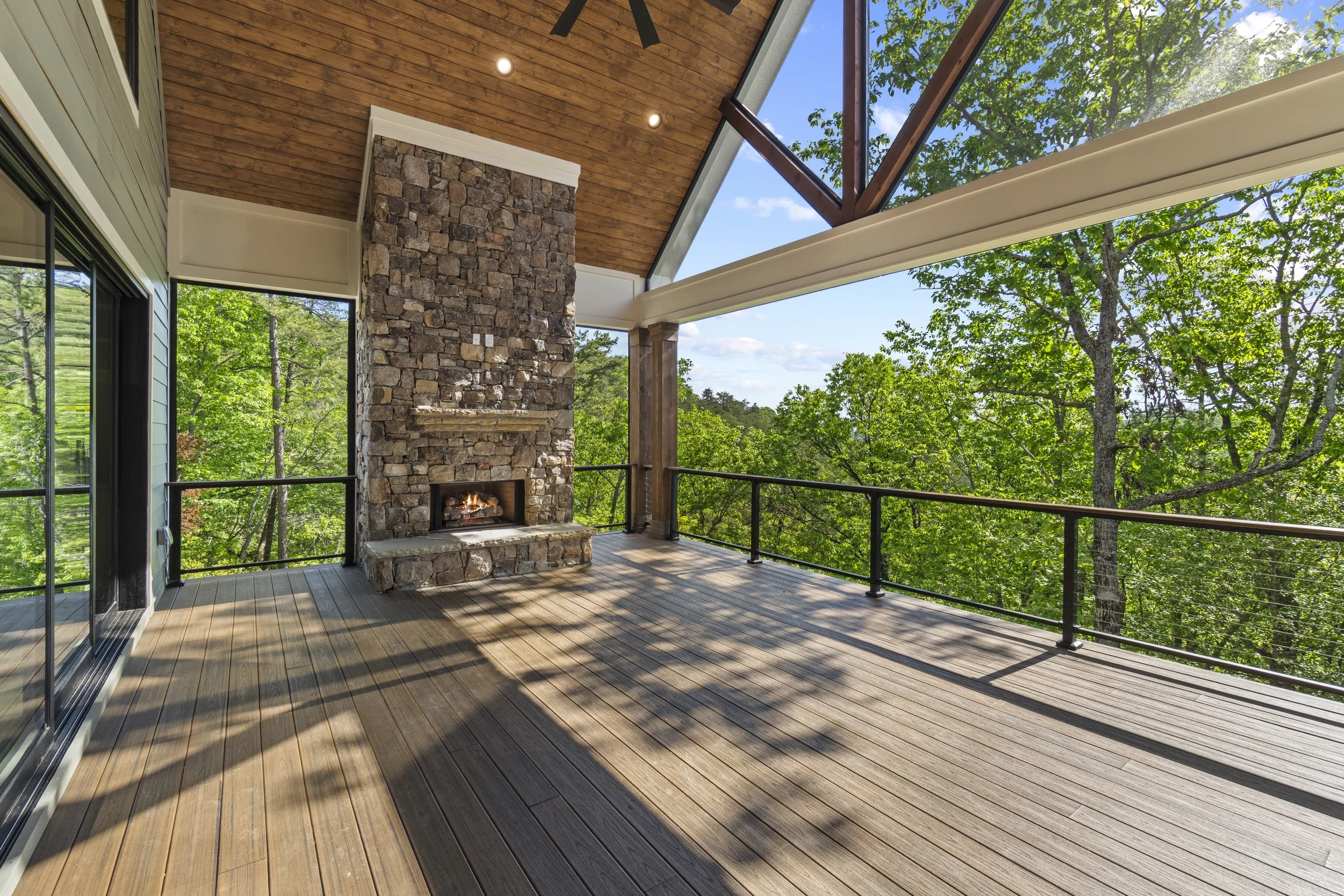A spacious outdoor balcony with wooden flooring, a stone fireplace, and a ceiling fan. The area is surrounded by green trees and features large glass doors and windows, with a partial view of the sky.