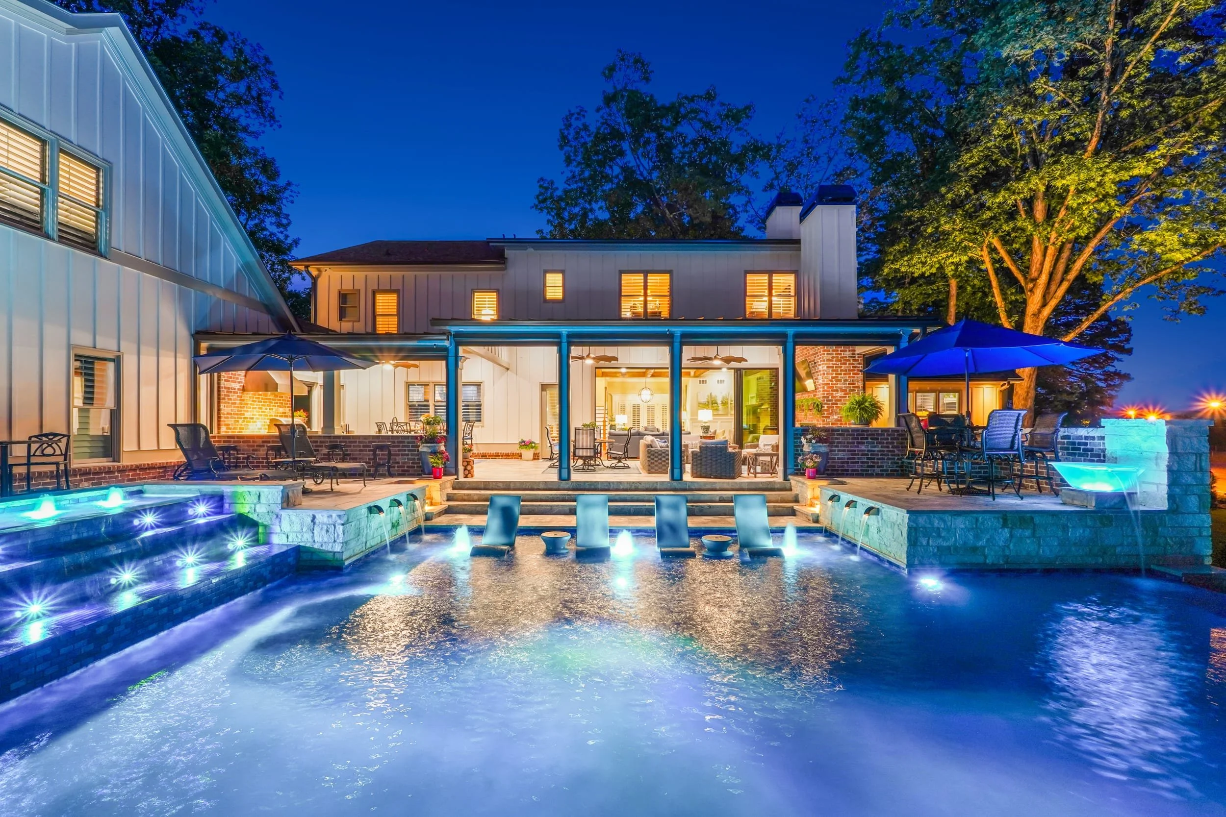 A backyard with a swimming pool illuminated with underwater lights at night. There is outdoor furniture with umbrellas on a patio, and a house with lit windows in the background.