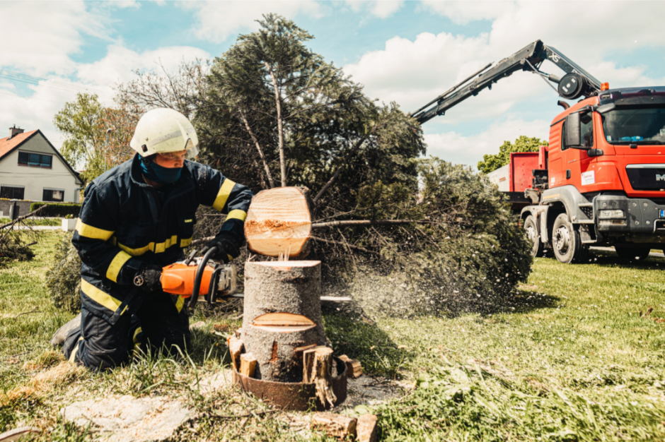 Firefighter using a chainsaw to cut a fallen tree in a grassy yard with a rescue truck in the background.
