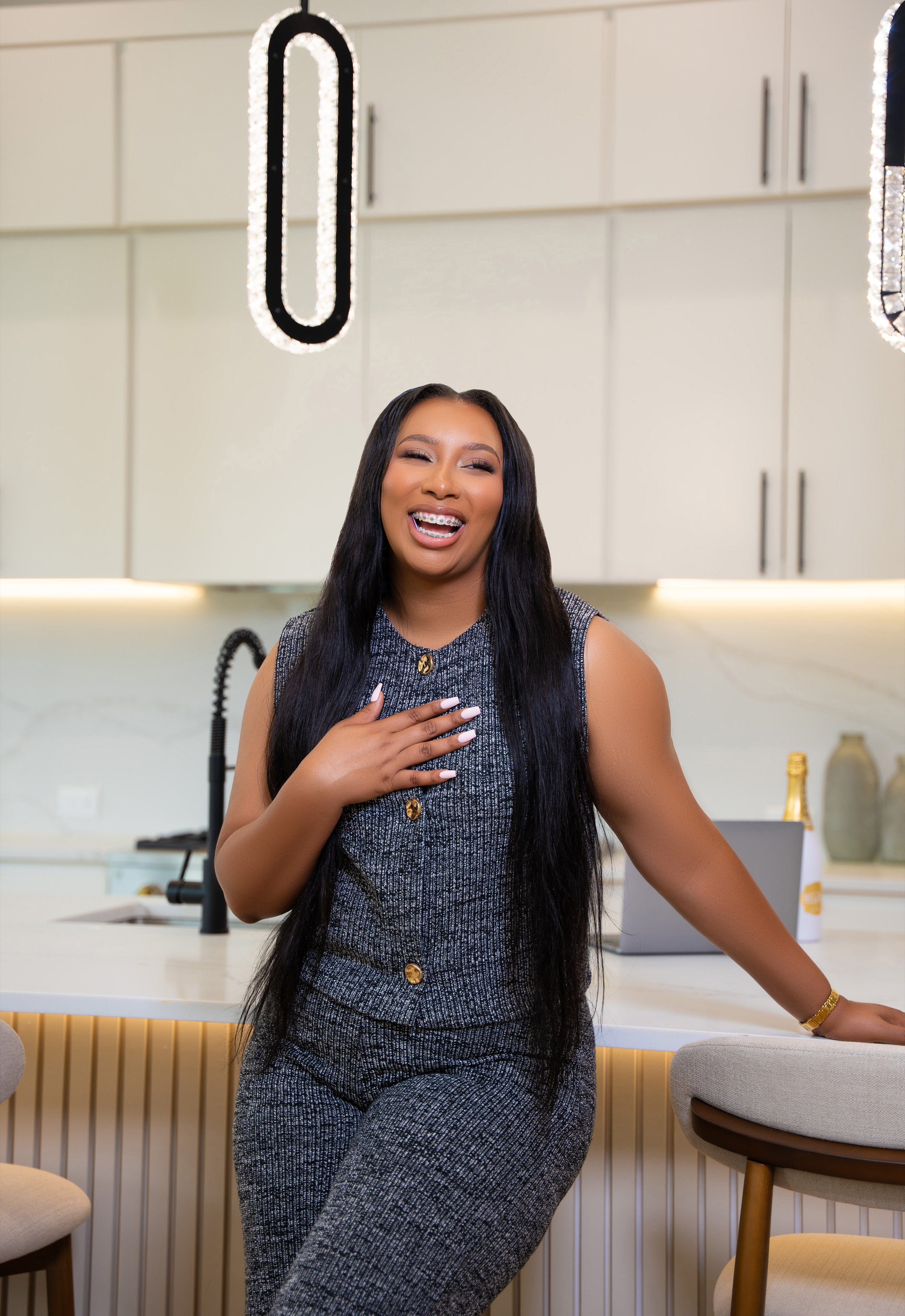A woman with long black hair, wearing a sleeveless gray patterned outfit, laughing and smiling in a modern kitchen with white cabinets and a white island, illuminated by contemporary pendant lights.