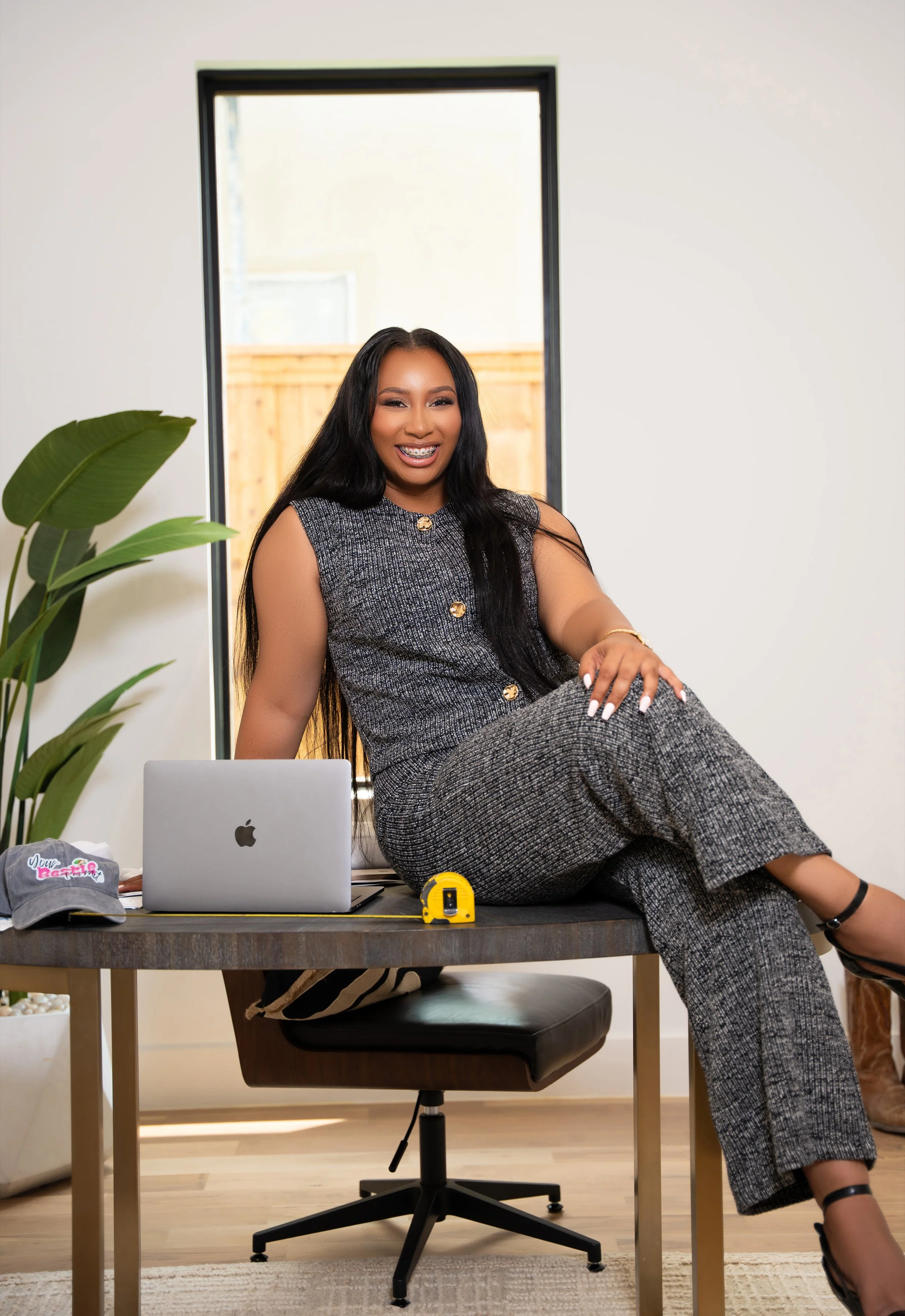 A woman in a patterned dress is sitting on a desk, smiling, with a laptop, a cap, and a tape measure on the desk, in a bright room with a plant and a window in the background.
