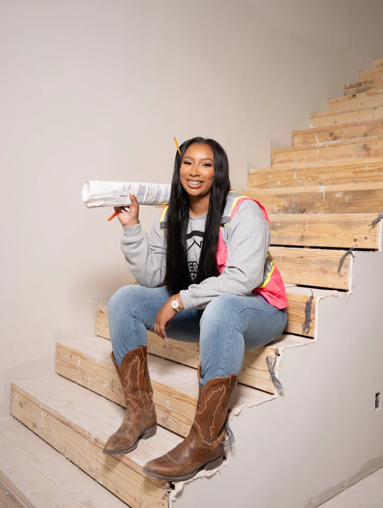 Woman in construction gear sitting on unfinished wooden staircase holding rolled blueprints, smiling.