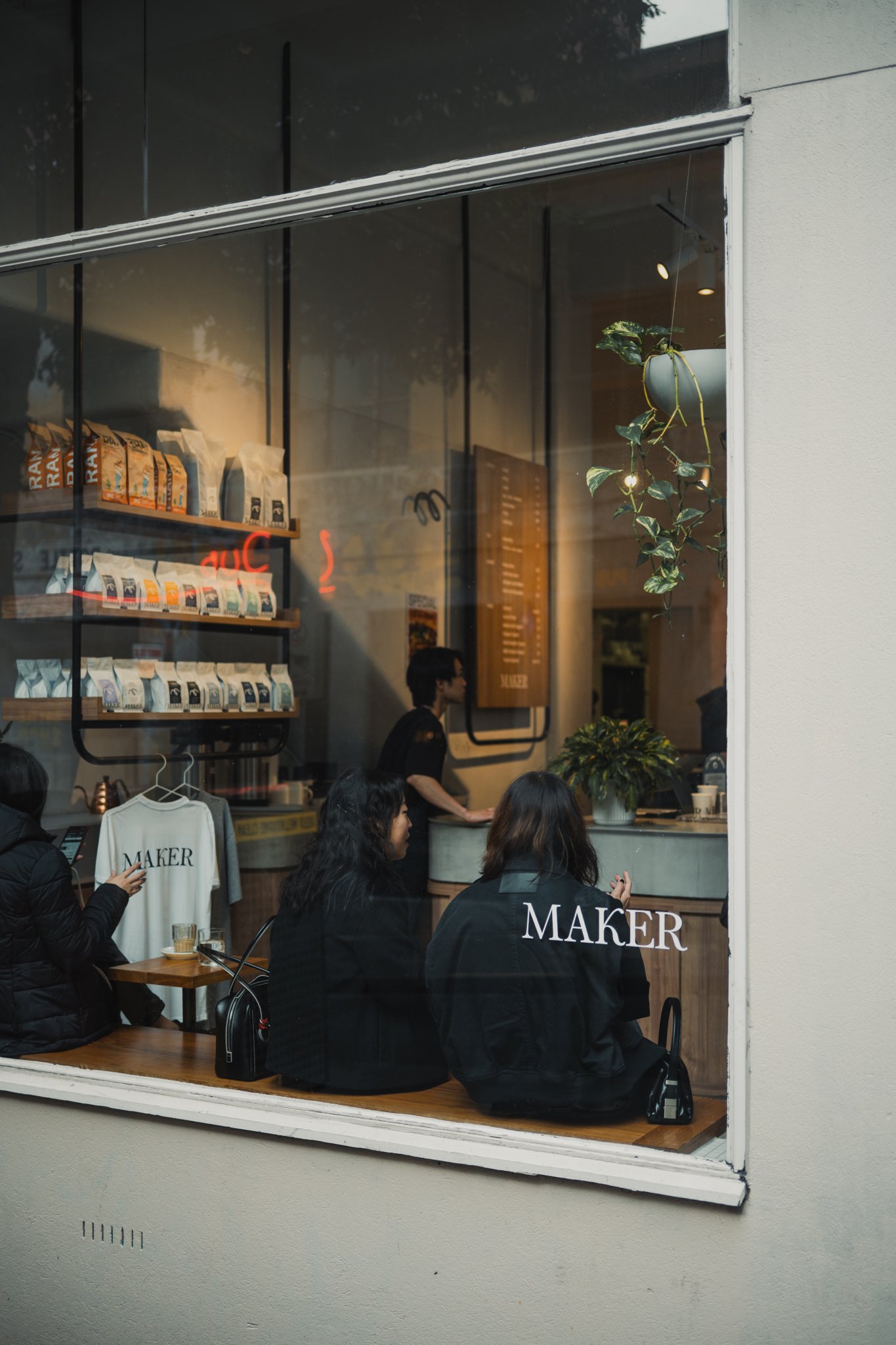 People sitting inside a coffee shop seen through a large window, with shelves of bags of coffee beans, a menu on the wall, and potted plants visible.