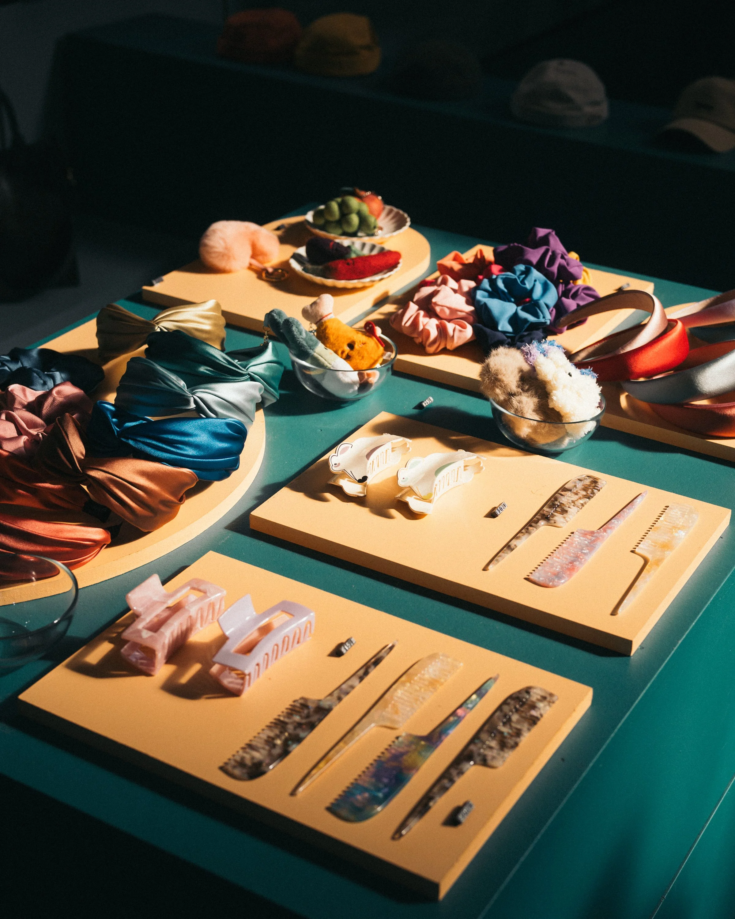 Display of colorful hair accessories, scrunchies, hair clips, and headbands on beige boards and glass bowls on a teal table.