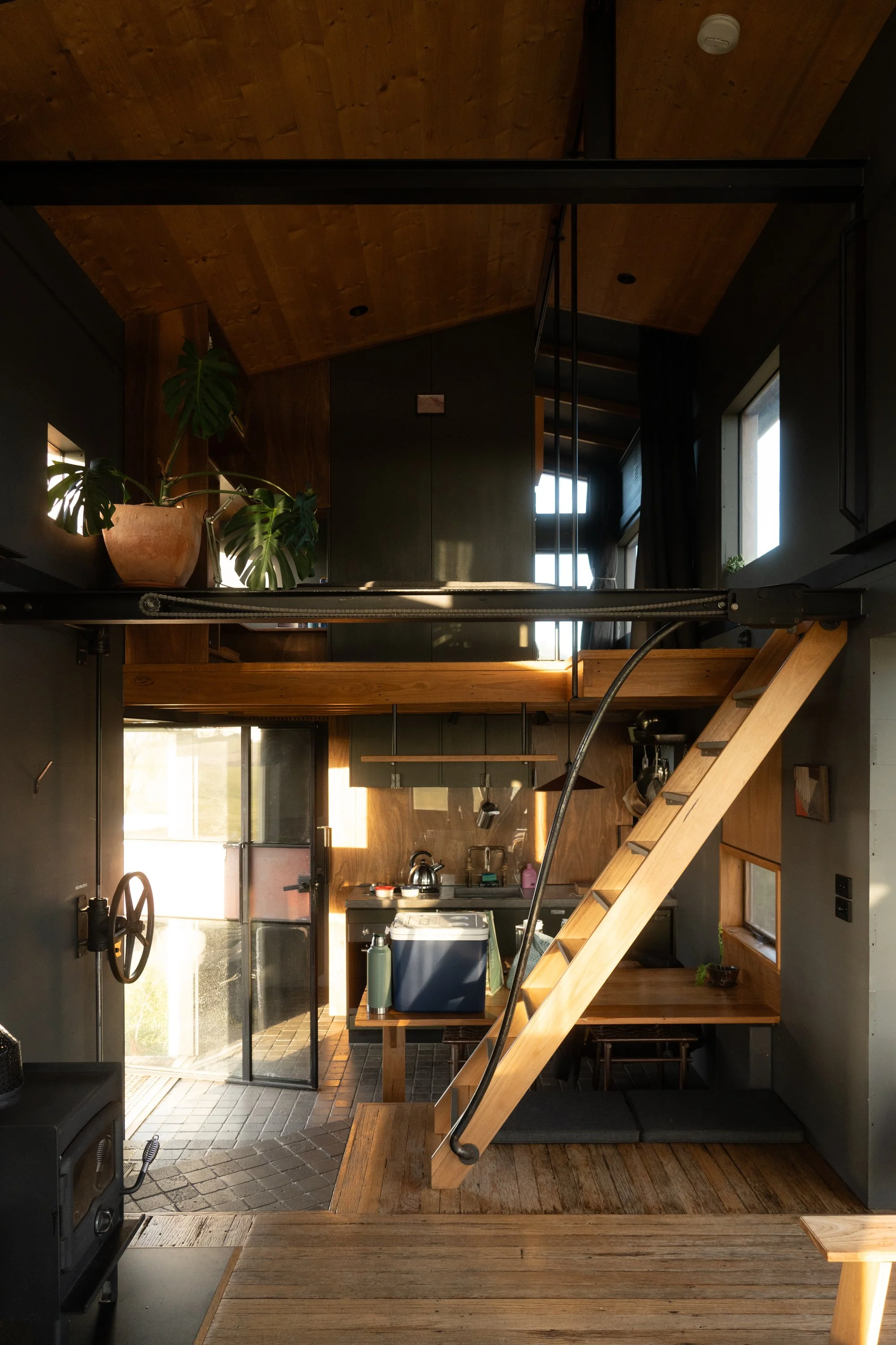 Interior of a small, modern apartment with a loft-style design, featuring wooden floors and loft stairs, a kitchen area with a countertop and appliances, and a potted plant by a window.