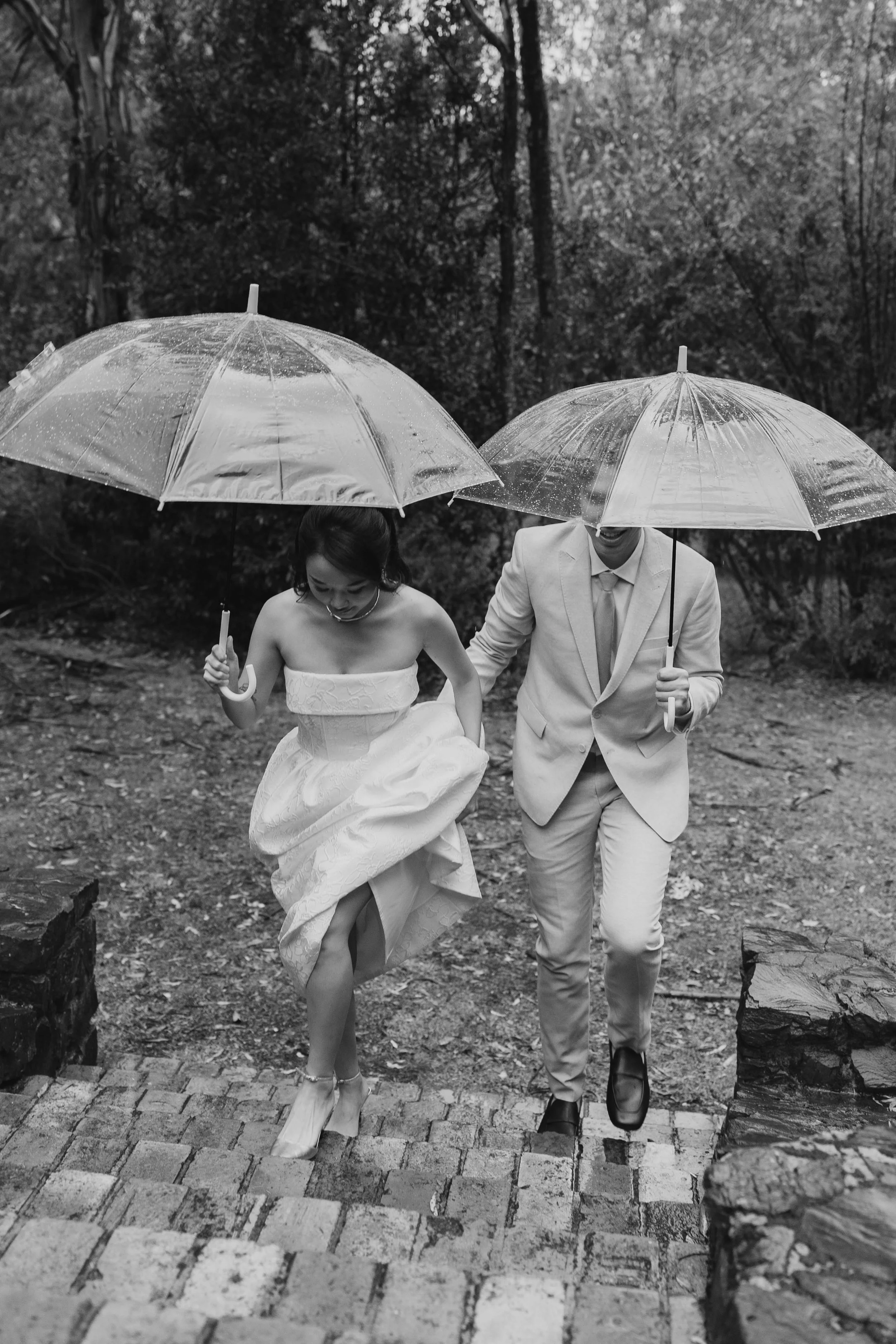 A black and white photo of a couple dressed in wedding attire walking on a path in a wooded area, each holding an umbrella in the rain.