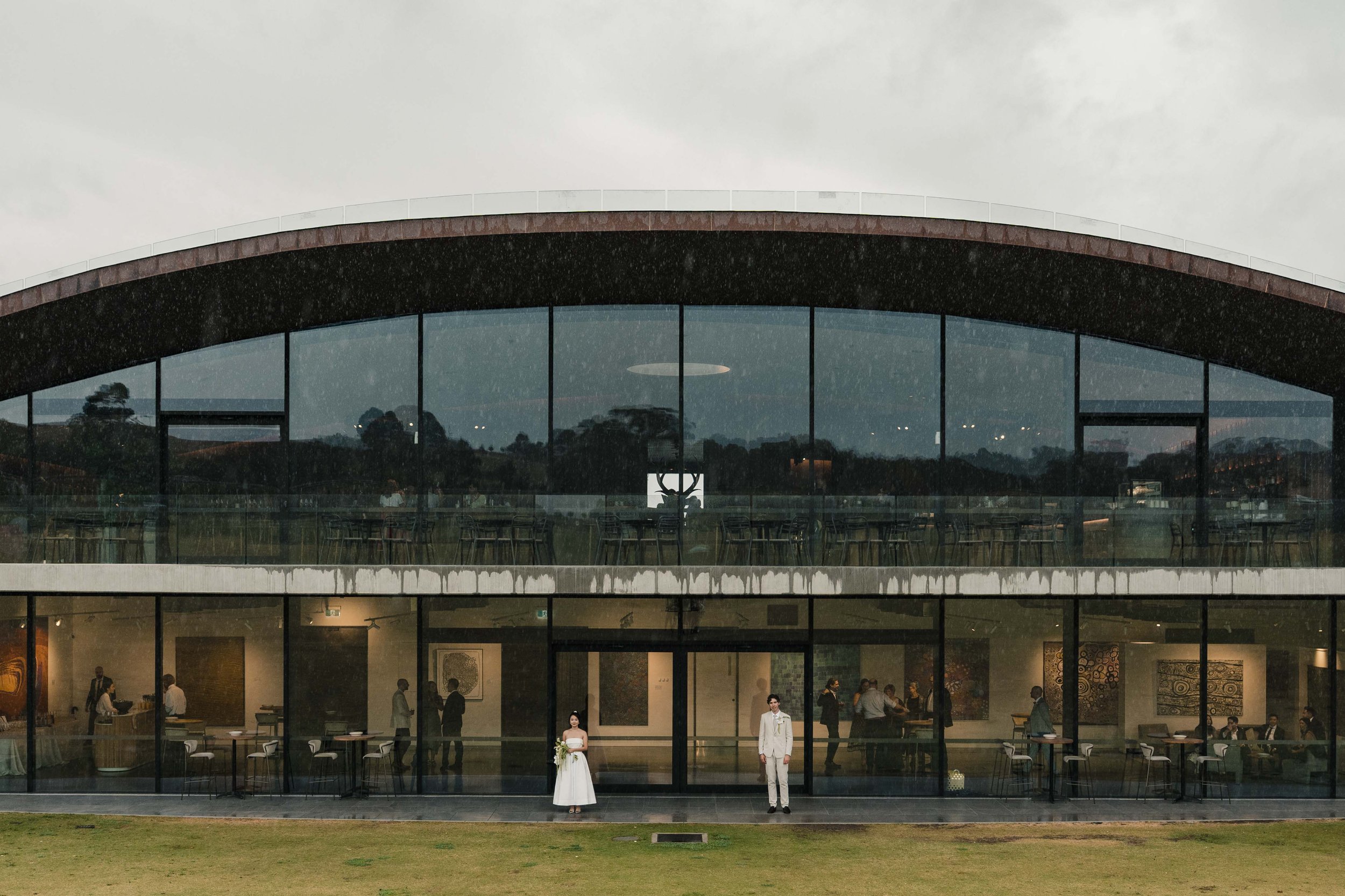 A modern two-story building with large glass windows, a curved roof, and a visible interior with people, including a couple dressed in wedding attire standing outside on the lawn.