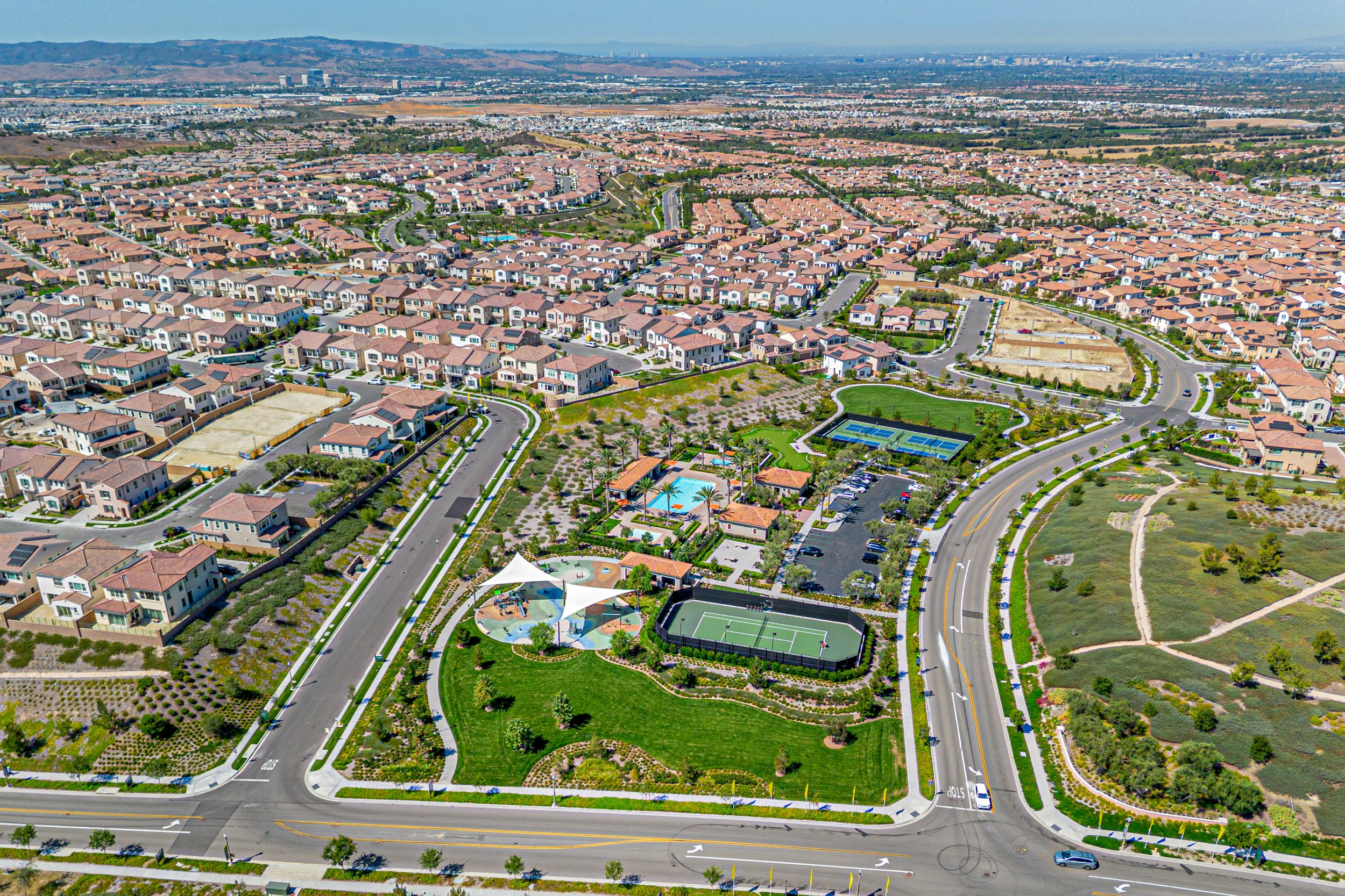 Aerial view of a suburban neighborhood with rows of houses, streets, a playground with tennis courts, and some open green spaces, set against distant mountains and a city skyline.
