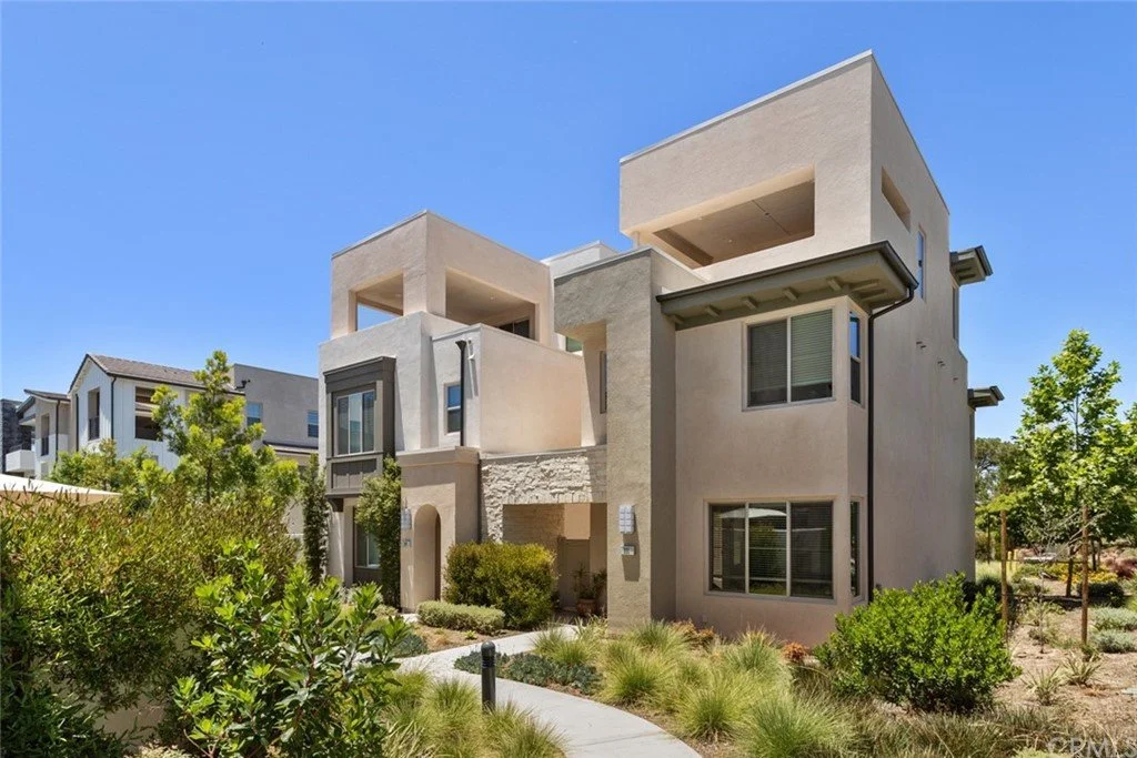Modern multi-story residential building with stucco exterior, balconies, and landscaped front yard under a clear blue sky.