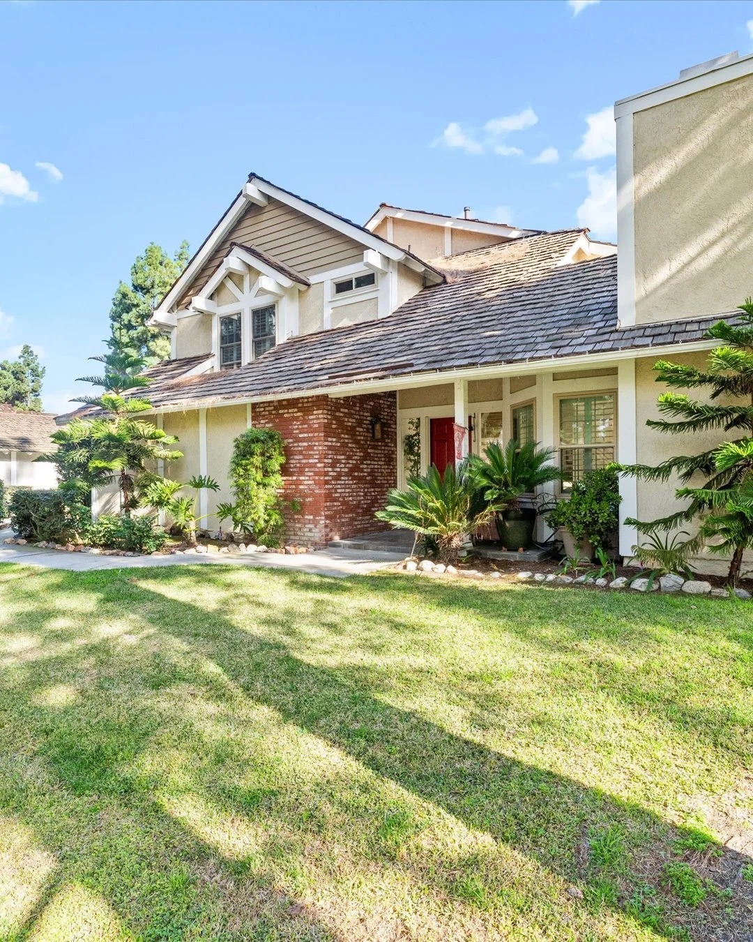 Front view of a two-story house with a brick and beige exterior, surrounded by a green lawn and plants, under a blue sky with some clouds.