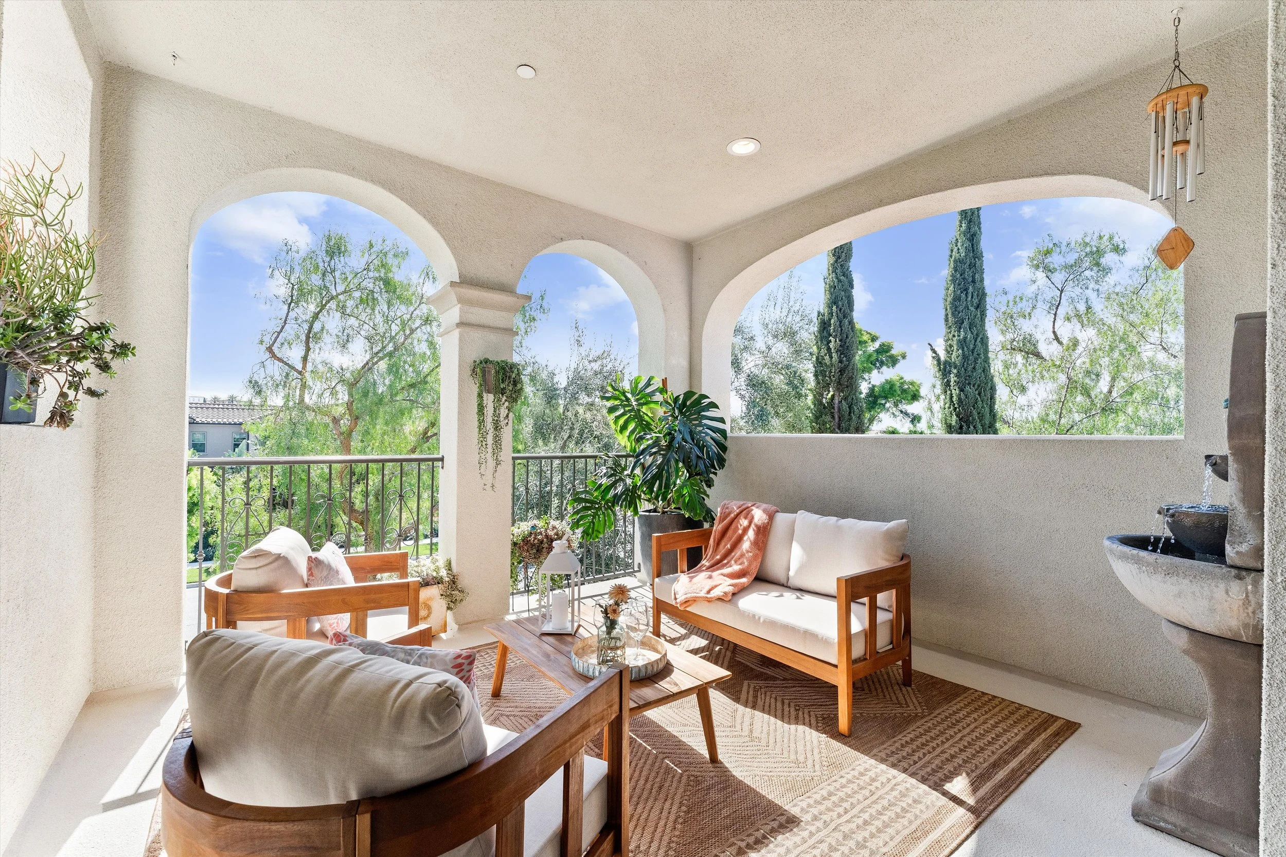 A balcony with outdoor seating, including two wooden chairs with white cushions and a small wooden table with decorative items, surrounded by potted plants and overlooking lush green trees.