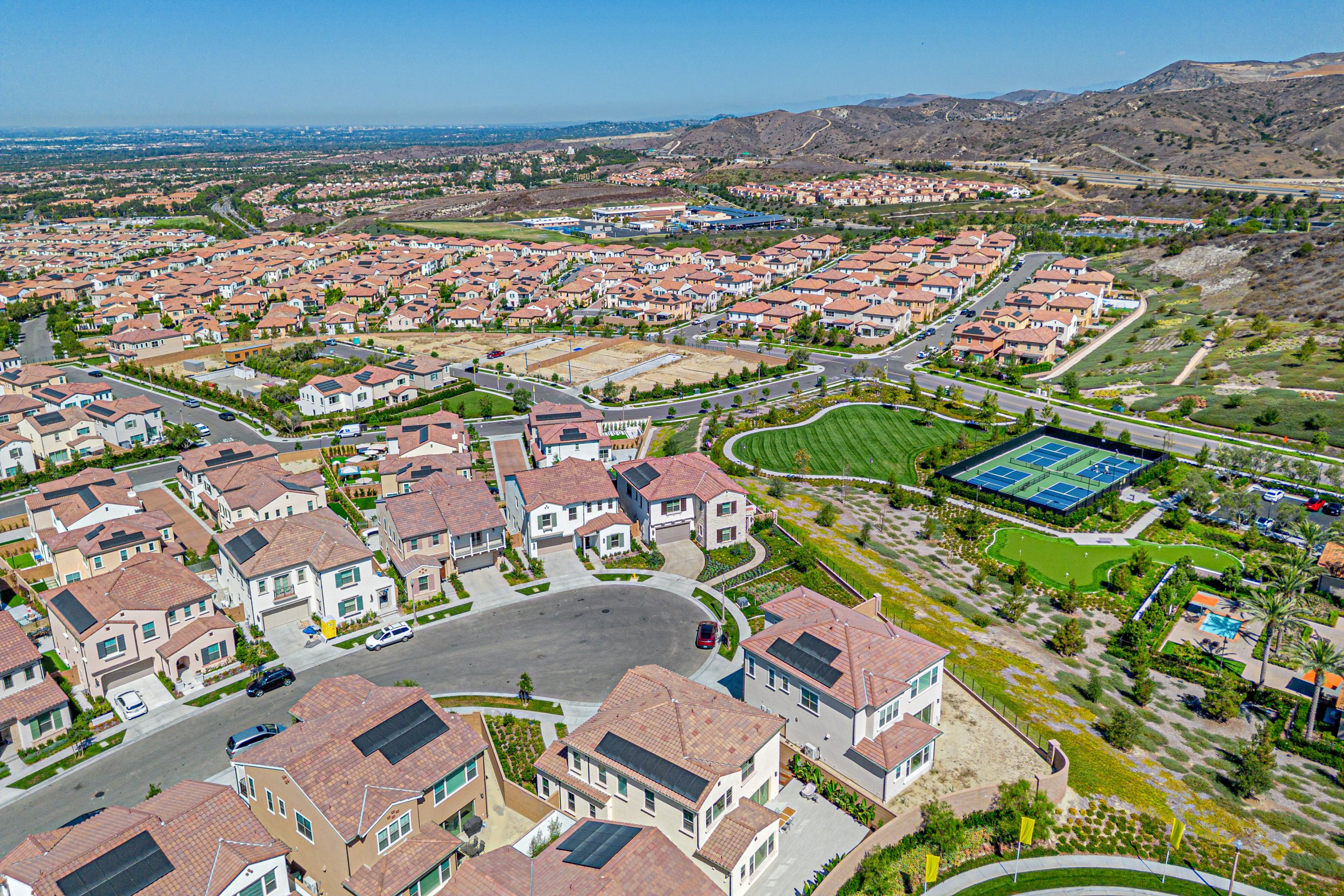 Aerial view of a suburban neighborhood with houses, streets, tennis courts, and a park, surrounded by hills in the background.