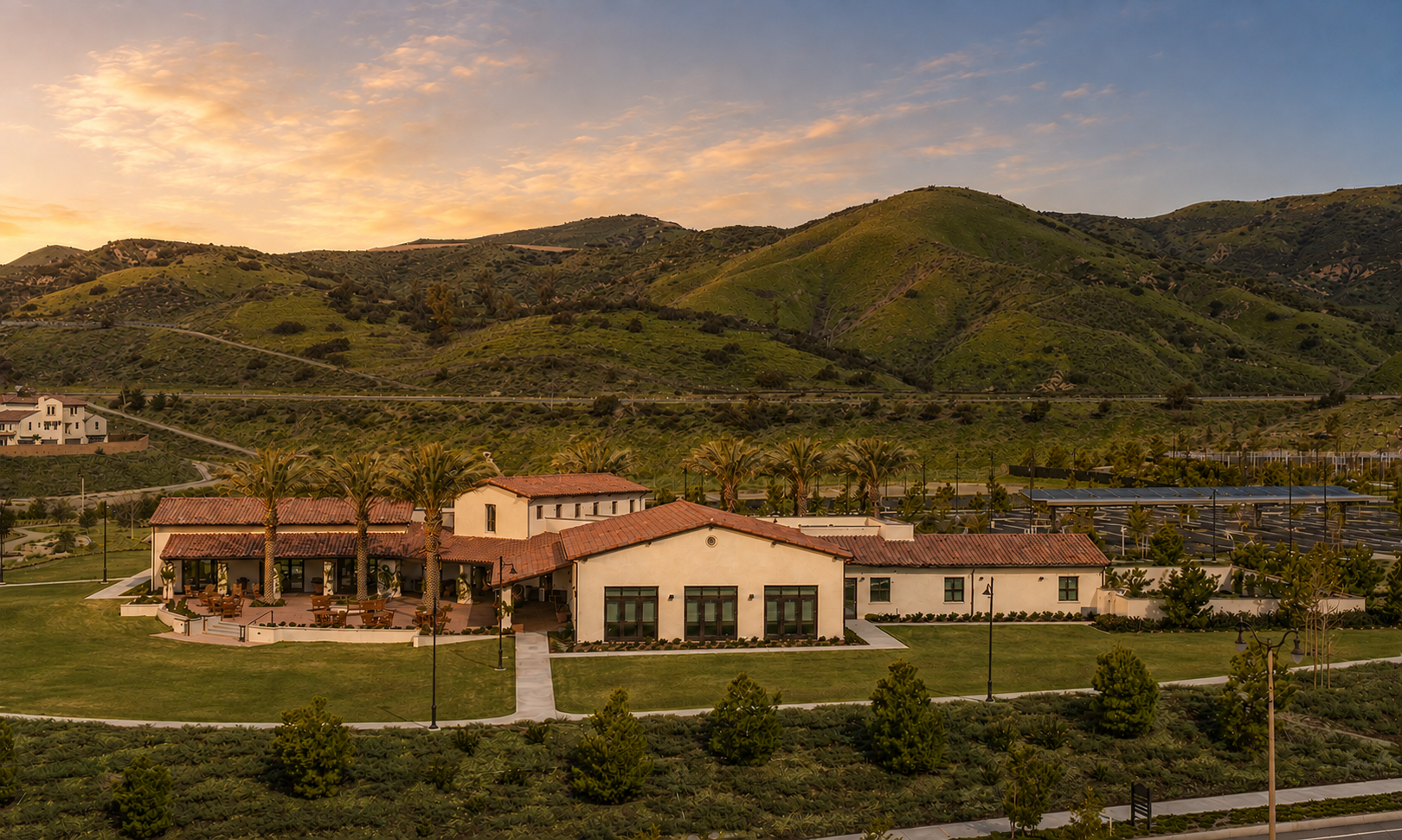 A scenic view of a building with red-tiled roof and palm trees, surrounded by lush green lawns and hilly landscape under a partly cloudy sky at sunset.