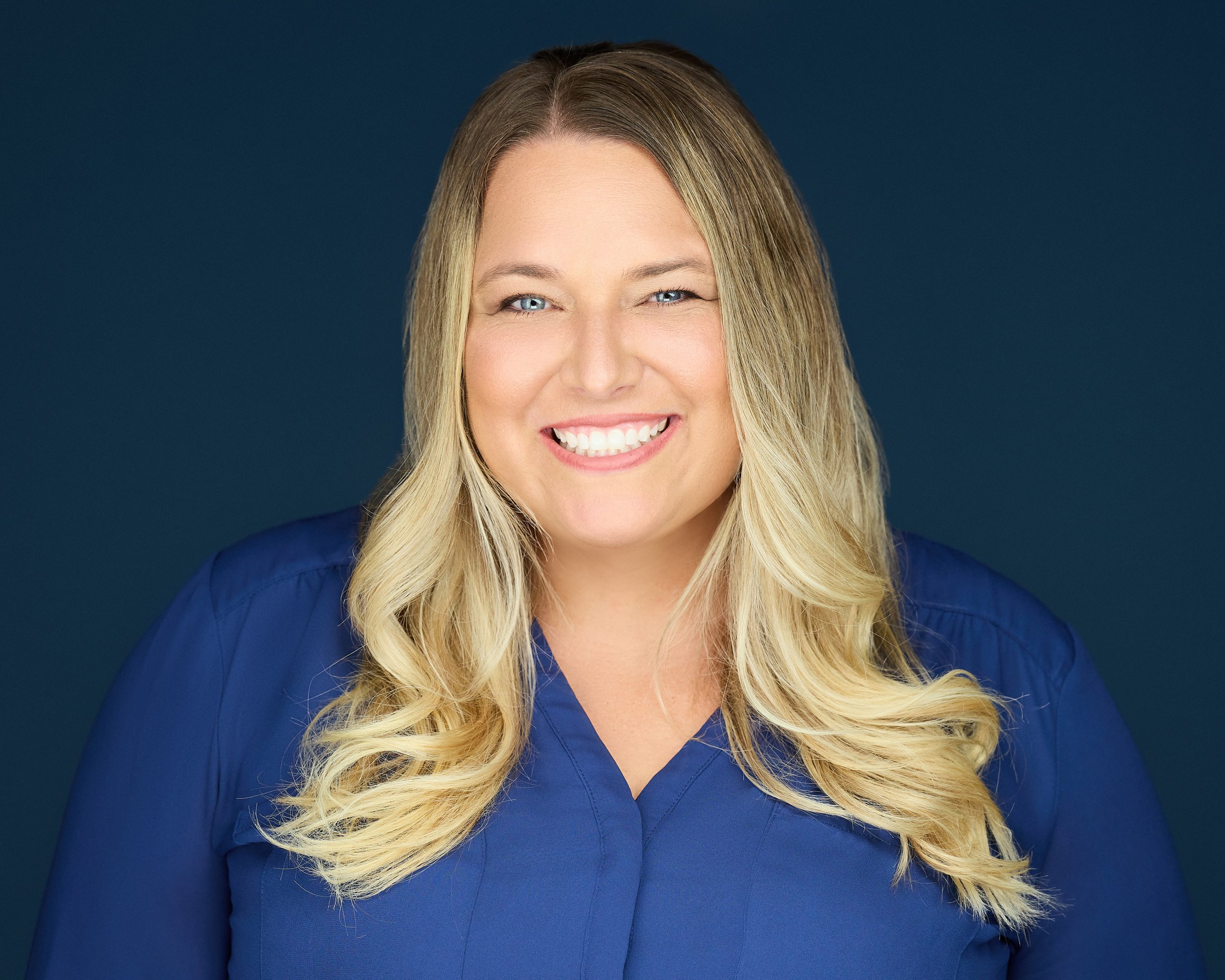 A smiling woman with long blonde hair and blue eyes, wearing a blue top, against a dark blue background.
