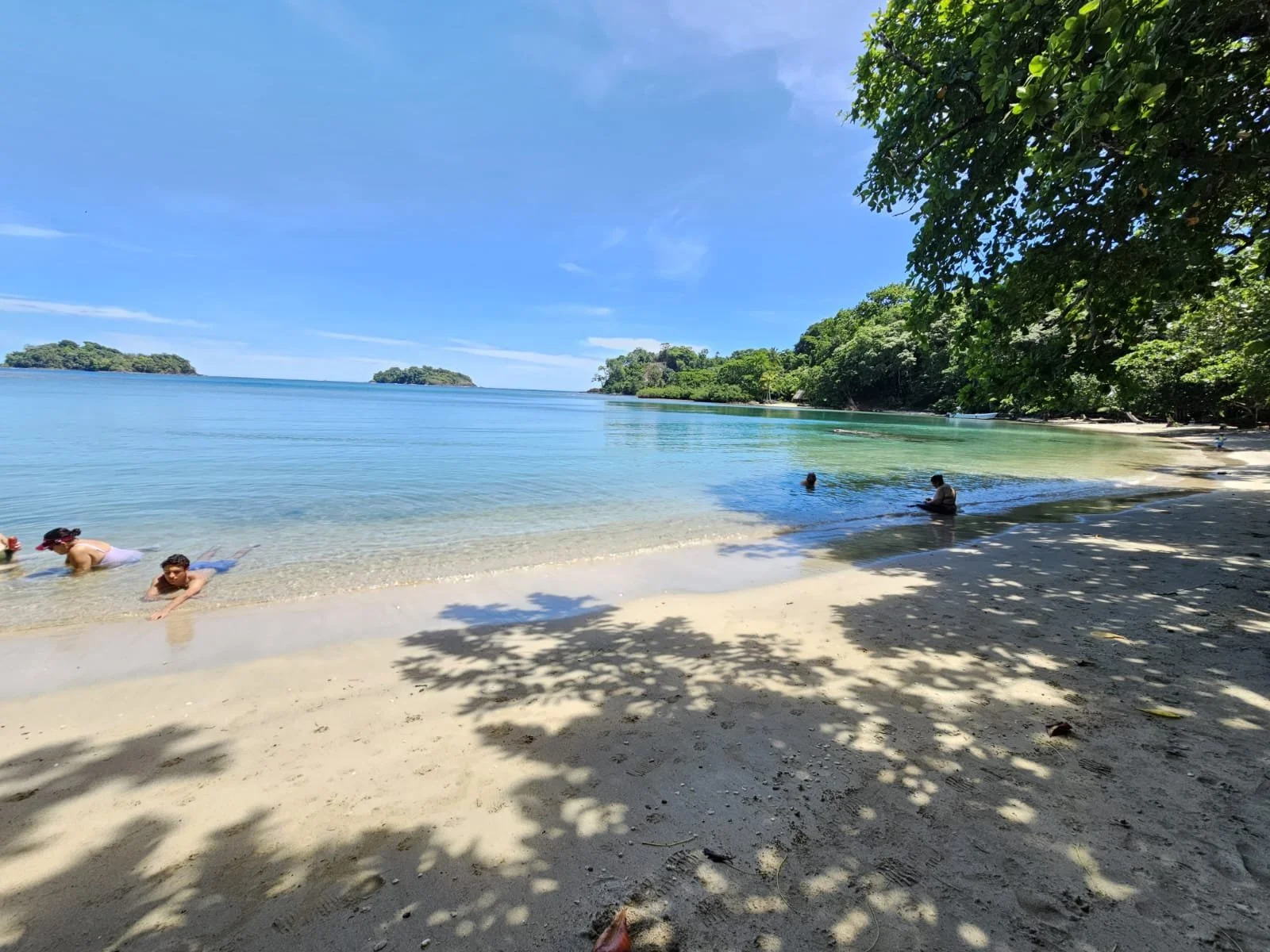 Playa tropical con arena blanca, aguas tranquilas y árboles frondosos con sombra; varias personas disfrutando en el mar, algunos bañándose y otros descansando en la orilla; pequeñas islas en el horizonte bajo un cielo azul con pocas nubes.