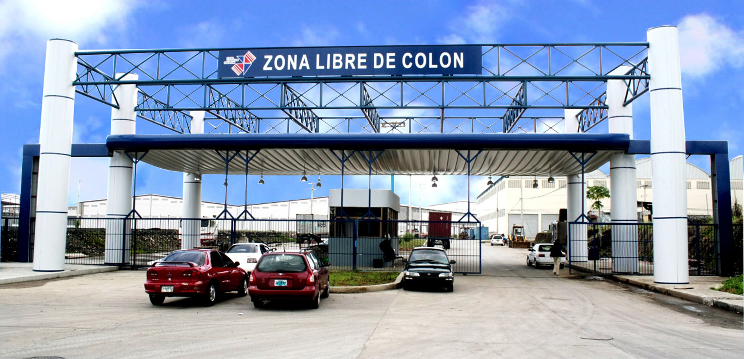 Entrada a una zona libre de colon con un cartel que dice 'Zona Libre de Colon', varias autos estacionados y edificios en el fondo.