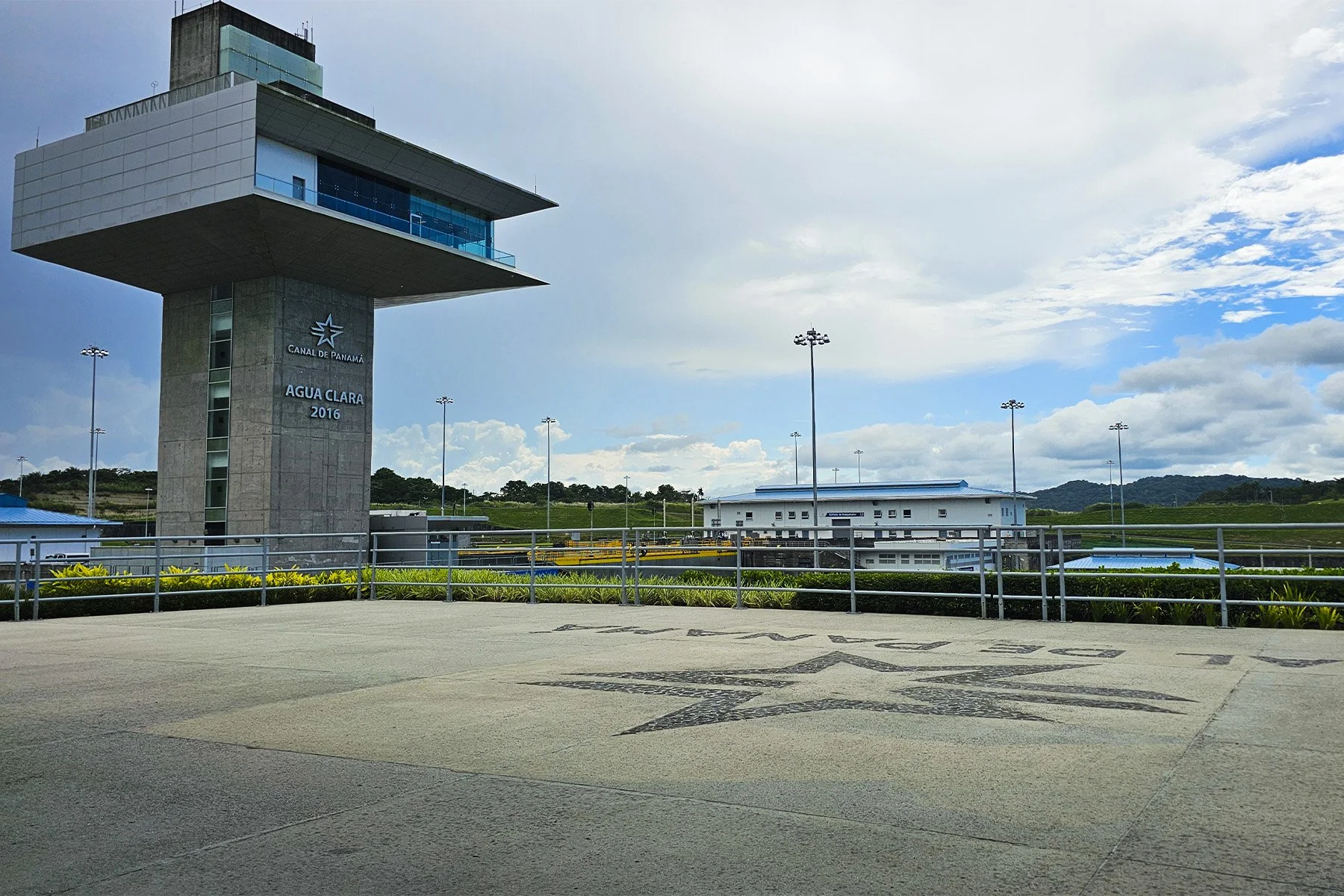 Vista de una torre de control en un aeropuerto con cielo parcialmente nublado, vegetación en el fondo y señalización en el piso.