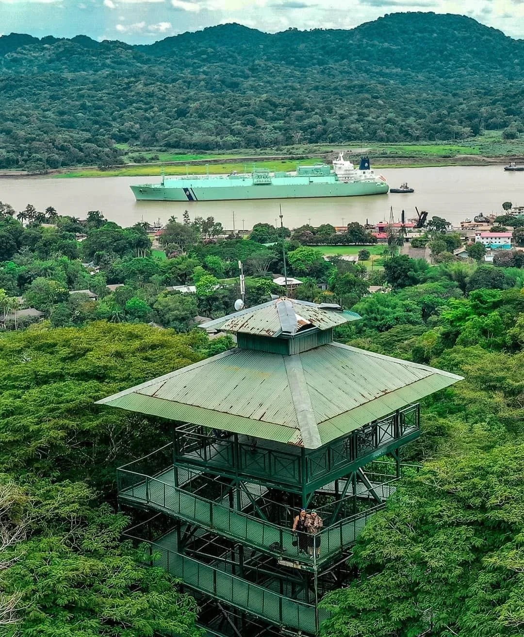 Vista aérea de un área natural con un edificio en lo alto de los árboles, un río y un buque de guerra en el agua, montañas en el fondo.
