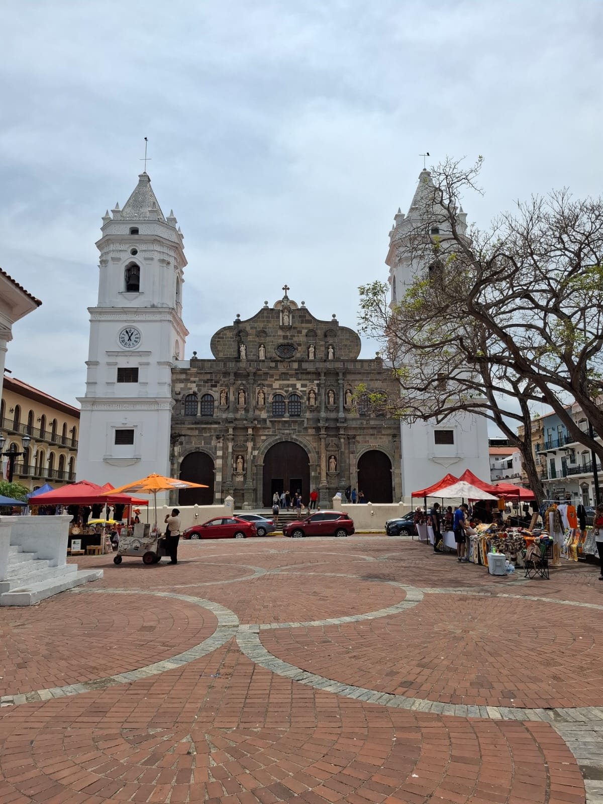 Iglesia con fachada de piedra y dos torres blancas, en una plaza con puestos de vendedores y árboles con pocas hojas.