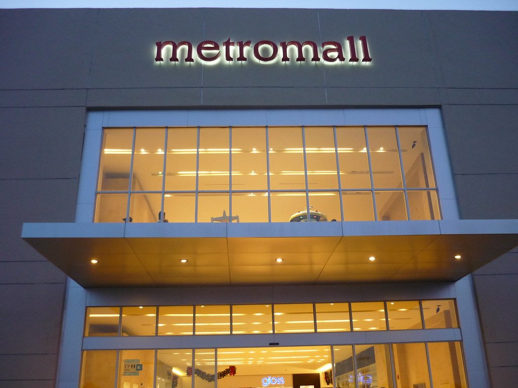 Vista frontal de la entrada de un centro comercial llamado Metromall, con ventanas grandes y iluminación interior visible.