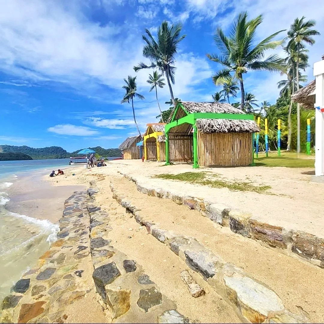 Cabañas coloridas en la playa con palmeras y un cielo azul con algunas nubes, personas en la arena y un bote en el agua.