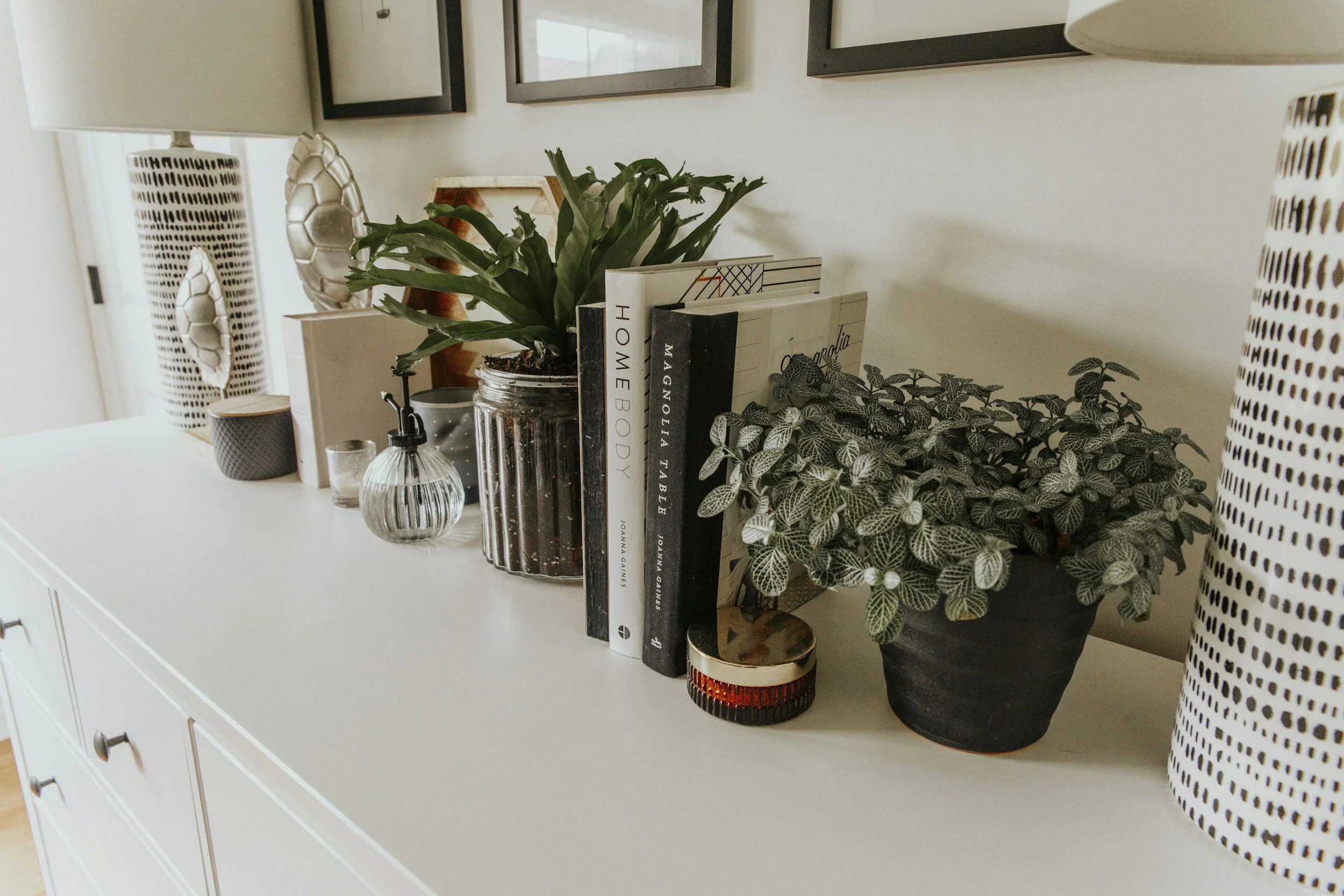 A white sideboard with decorative plants, books, and small objects on top, against a wall with framed art and patterned curtains.