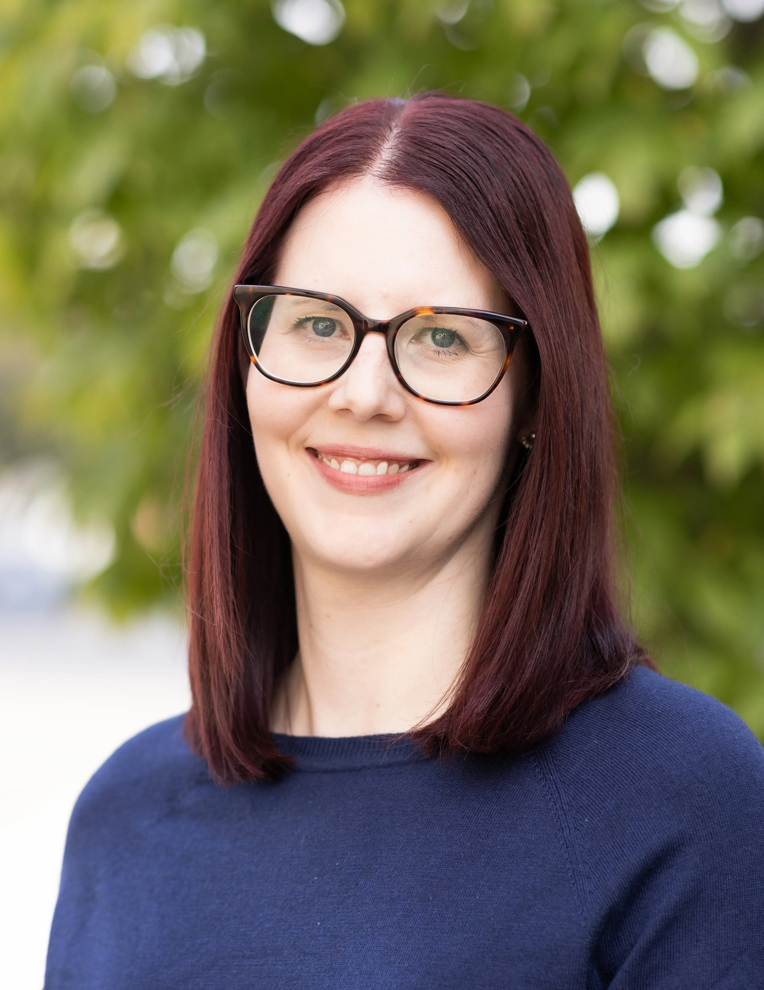 Close-up of Dawn with Auburn hair and glasses smiling, wearing a blue top, outdoors with a blurred tree background.