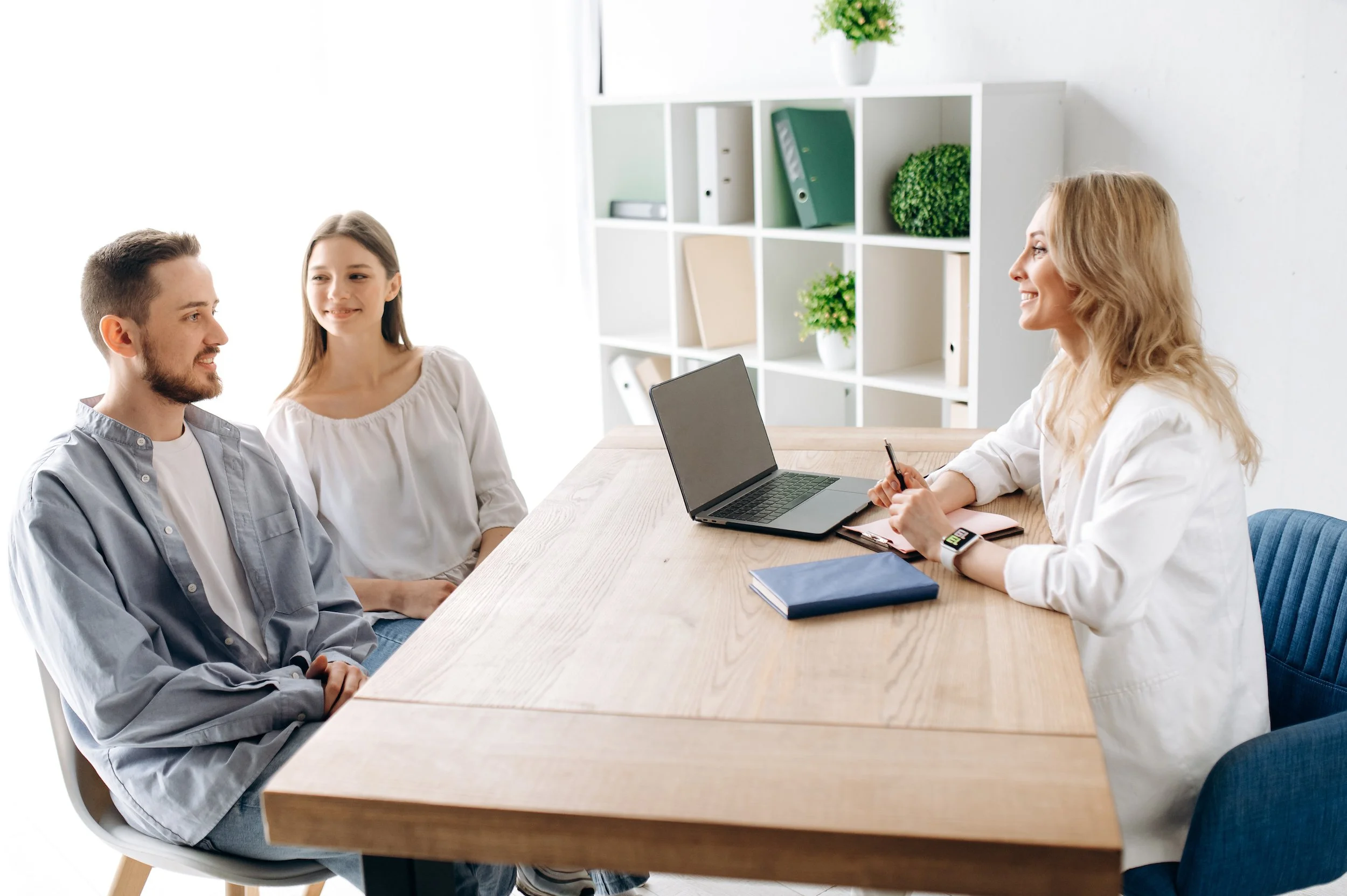 Three people sitting at a wooden table during a meeting in a bright, modern office. A man and a young girl are on one side, facing a woman with a laptop and notes. The woman is smiling, engaged in conversation with the other two.