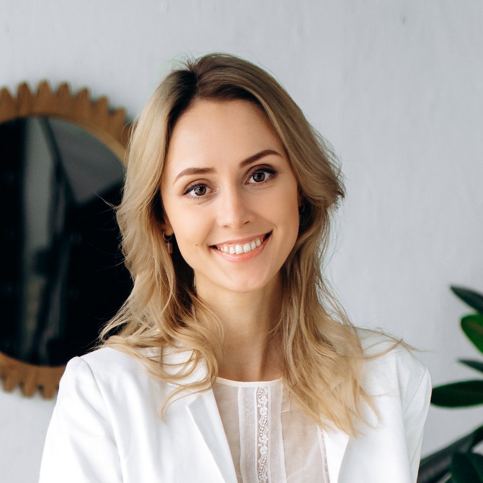 A young woman with blonde hair smiling at the camera, in a white top, in front of a round mirror and a green plant.