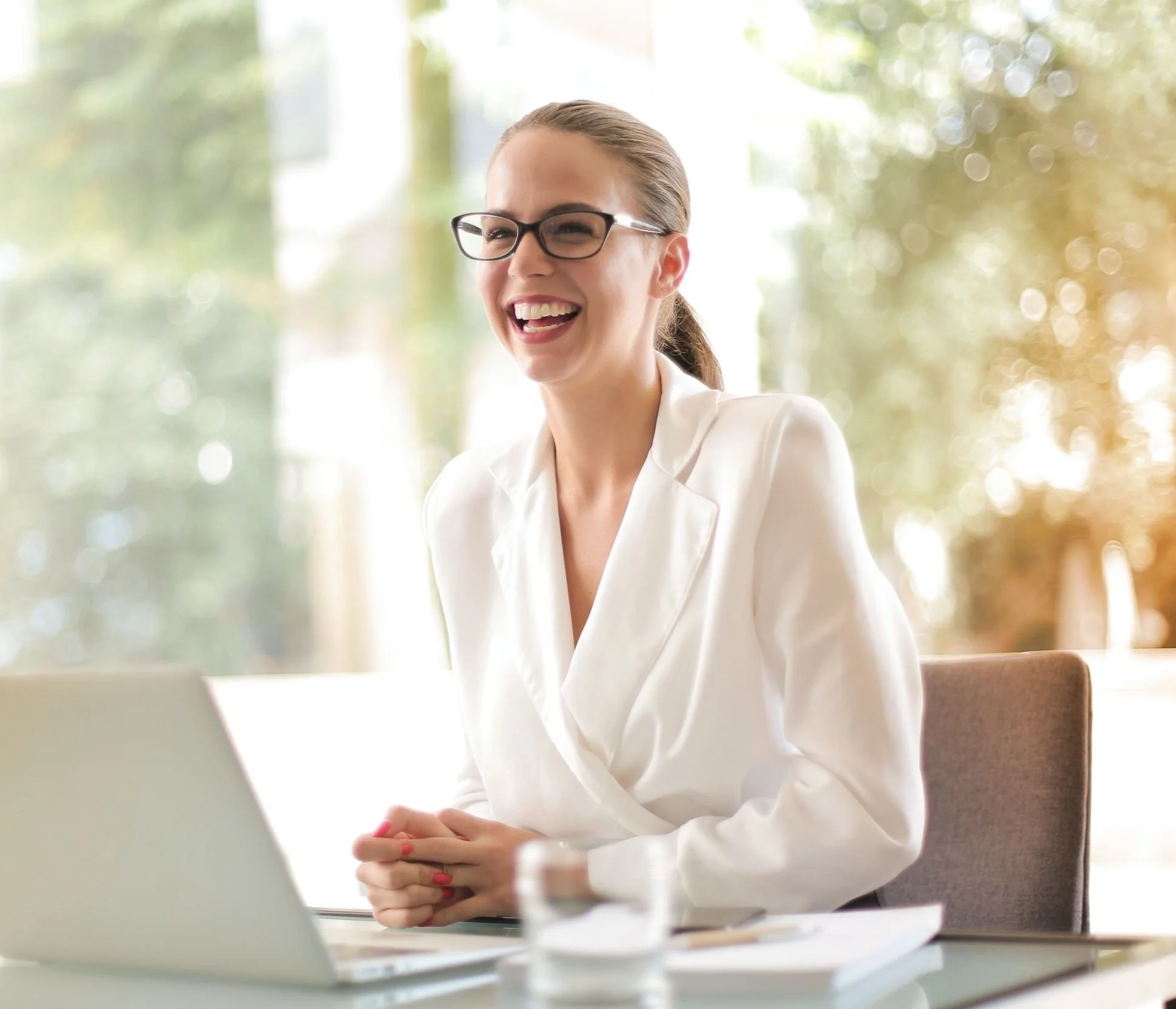 A woman with glasses and a white blazer sitting at a table, smiling and enjoying a moment during a meeting or conversation.