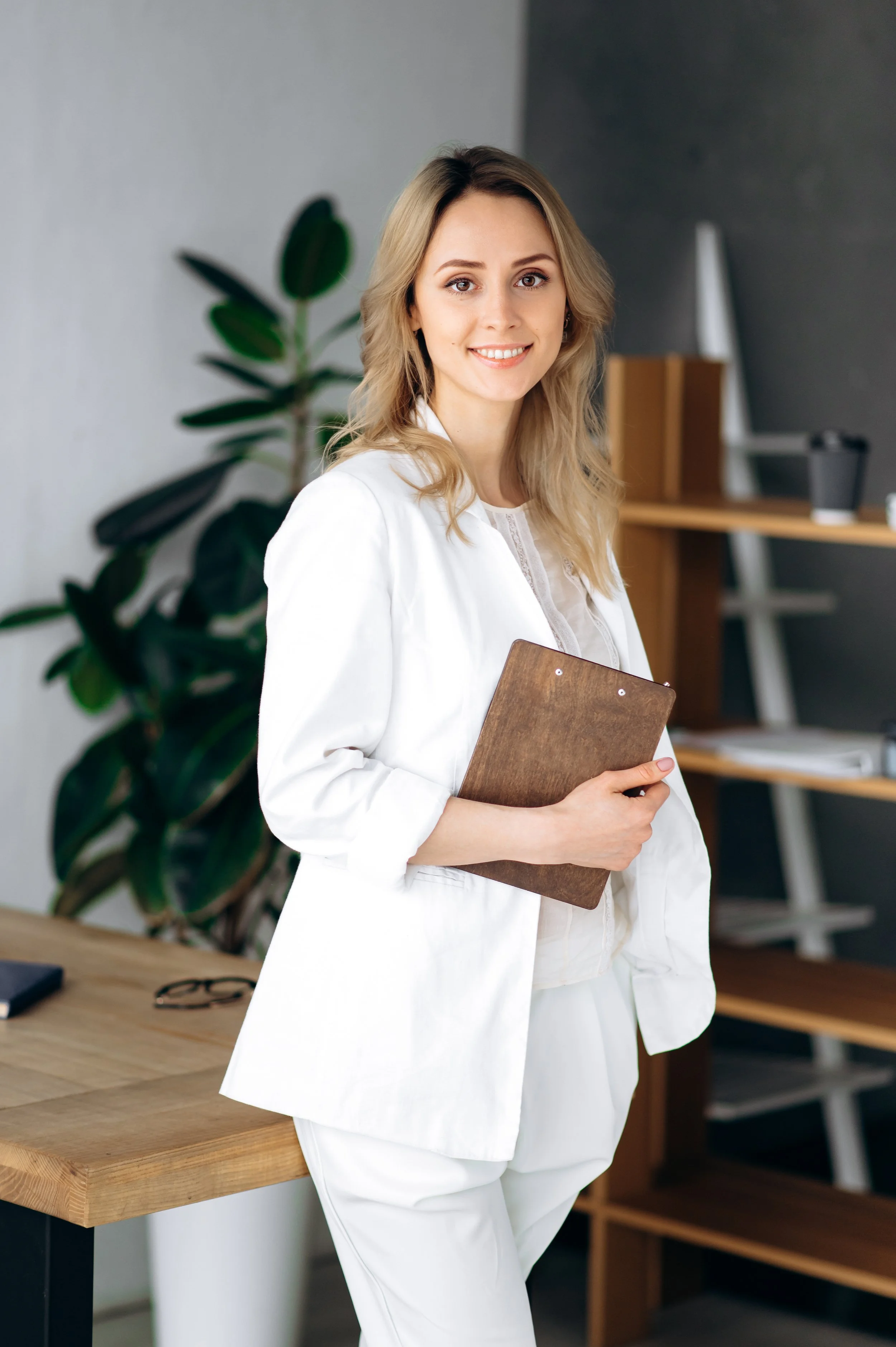 A woman with blonde hair, wearing a white blazer and holding a brown clipboard, stands in a modern office with a large green plant and shelves in the background.