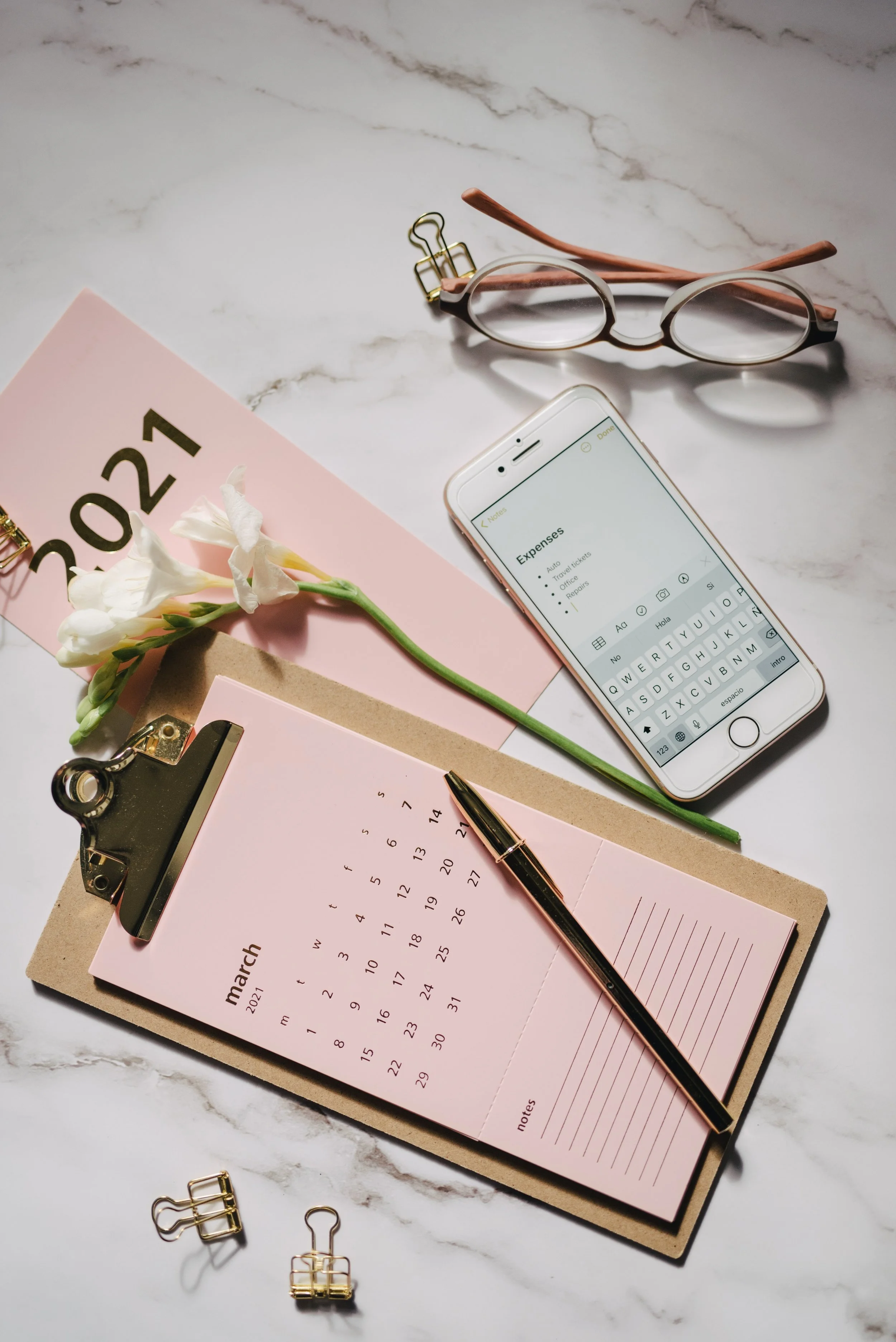 A desk setup with a pink March 2021 calendar, a white smartphone displaying expenses and notes, a black and gold pen, a small bouquet of white flowers, several gold binder clips, and pink eyeglasses on a white marble surface.