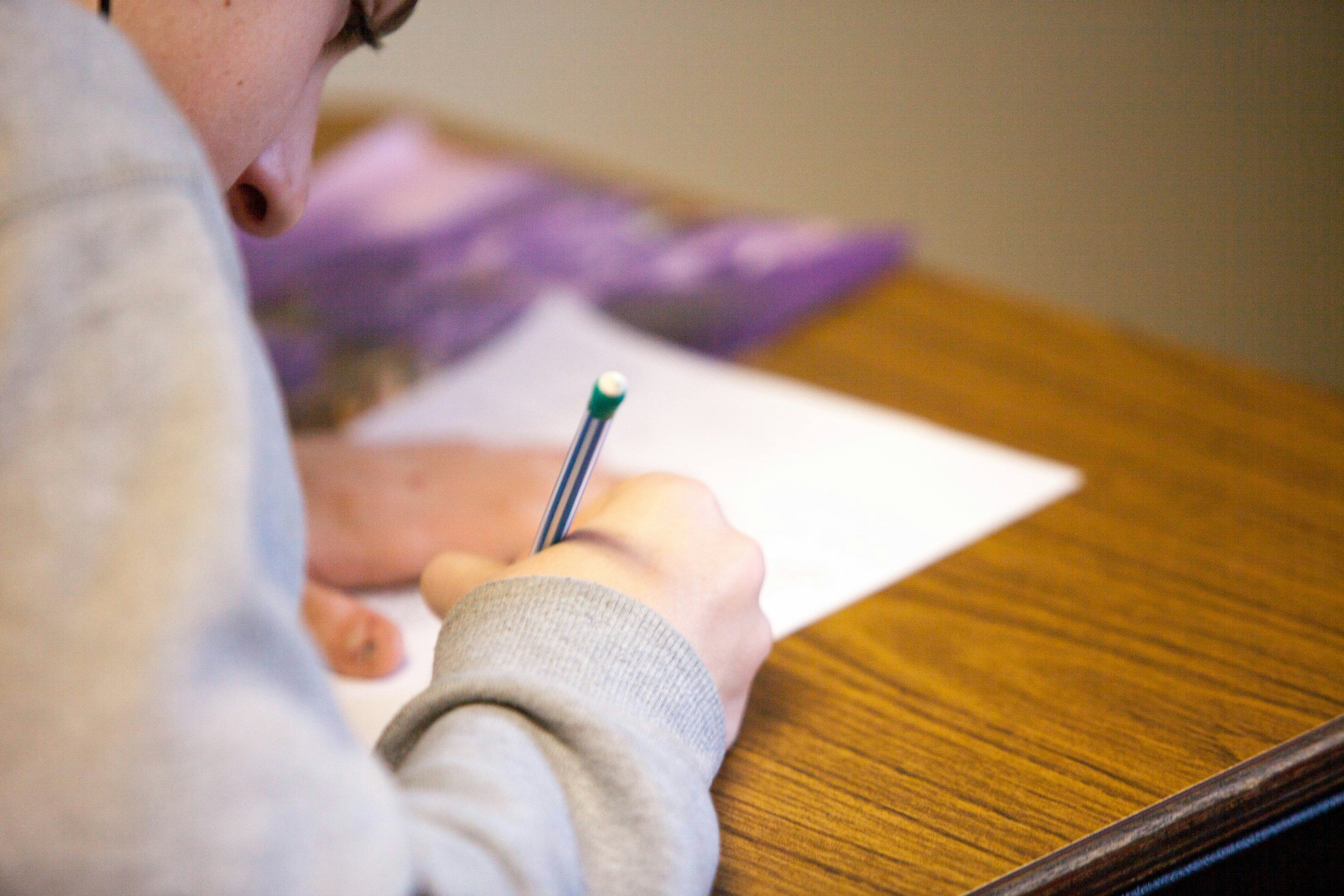 Close-up of a person writing with a pencil on a piece of paper, wearing a light-colored sweater, with a purple flower in the background.