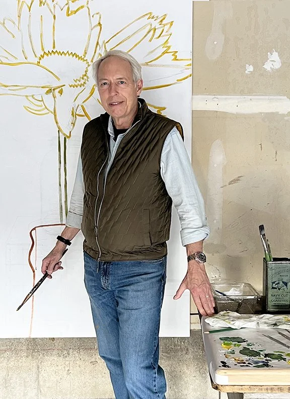 An older man standing in front of a large canvas with a yellow floral outline, holding a paintbrush and palette knife, wearing a brown quilted vest, white shirt, and jeans.
