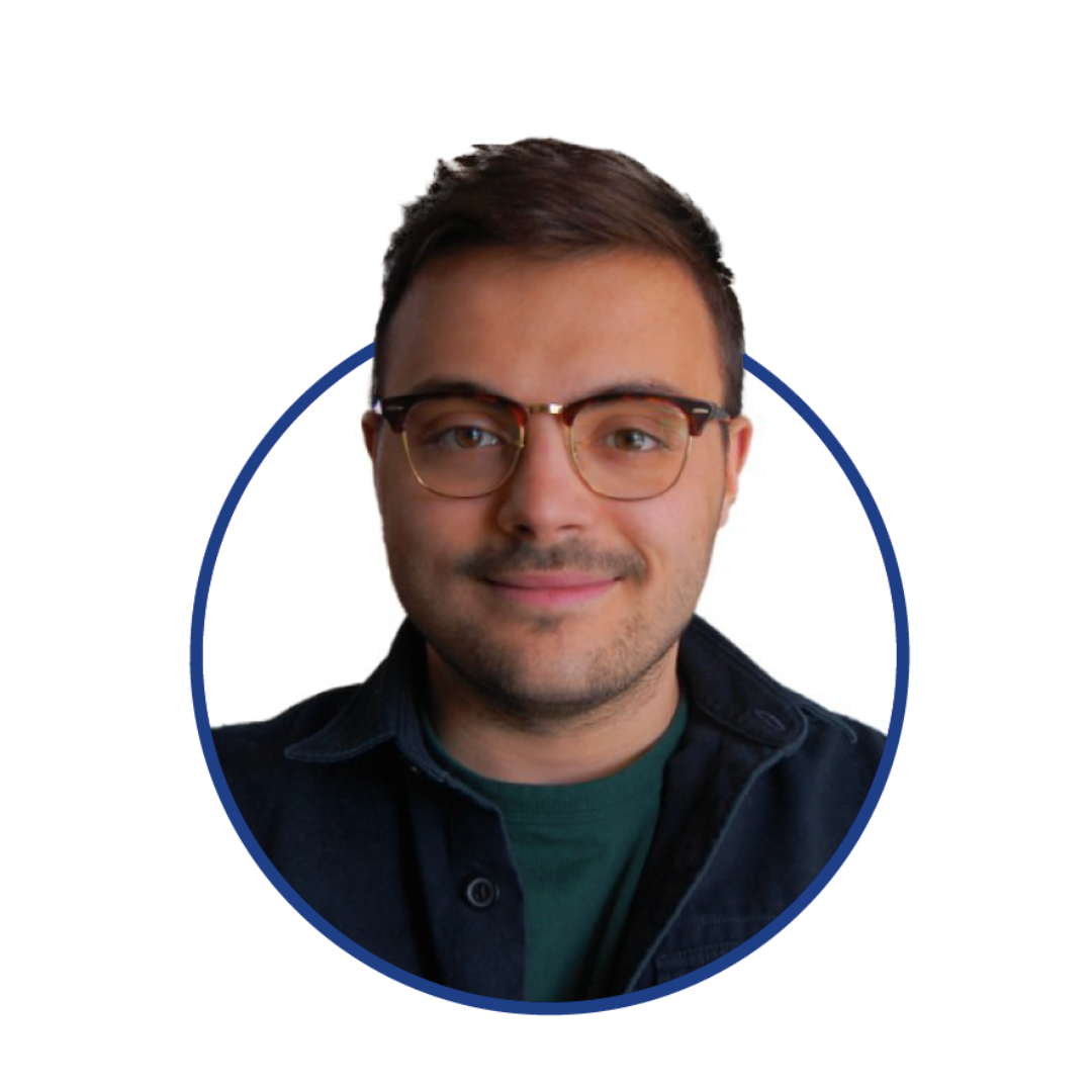 Headshot of a young man with short brown hair, glasses, a light beard, smiling, wearing a dark button-up shirt over a green t-shirt.