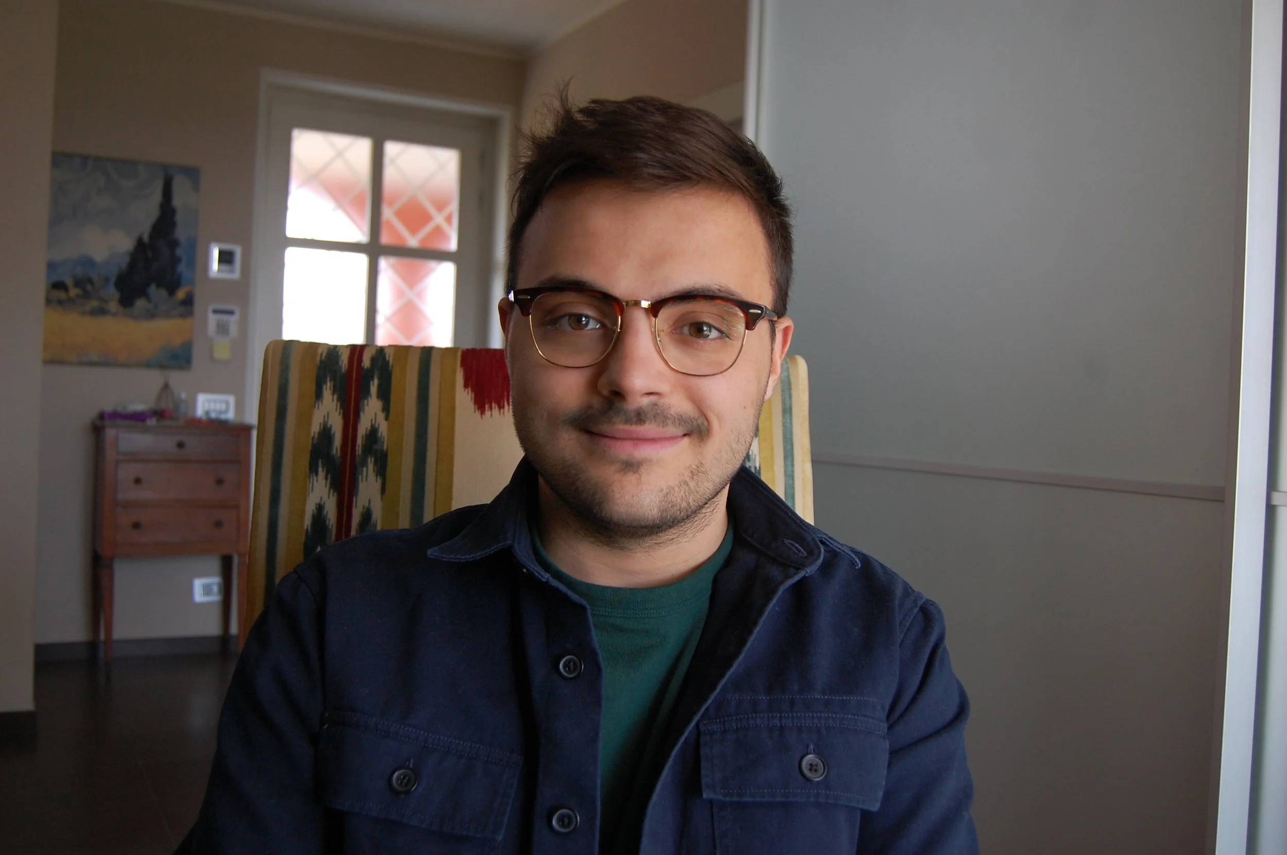 A young man wearing glasses and a dark jacket smiling while sitting indoors with a patterned chair behind him. In the background, there is a window, a painting, and a wooden dresser.