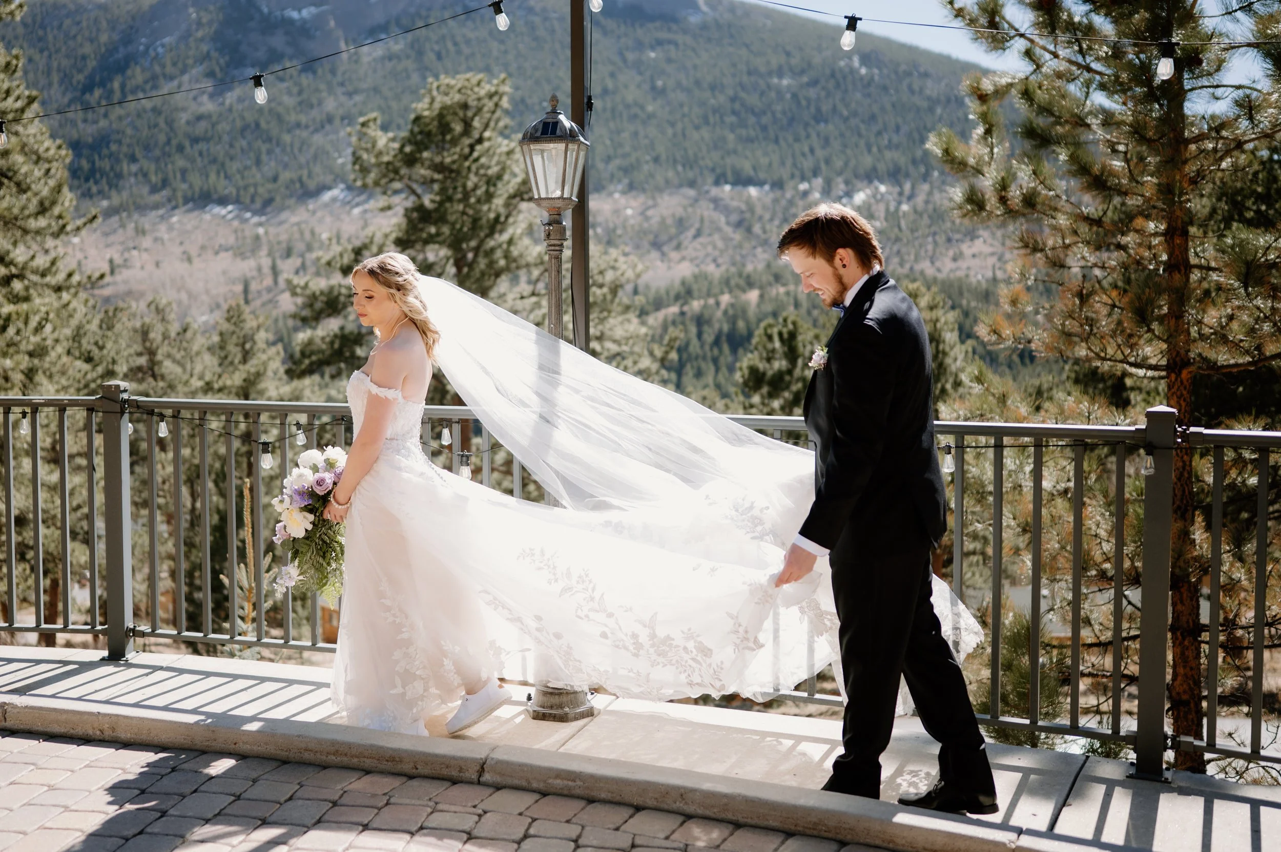 A married couple before their wedding at Della Terra in Estes Park Colorado, bride walking away carrying wedding bouquet of purple and white florals, groom holding train and veil in the wind