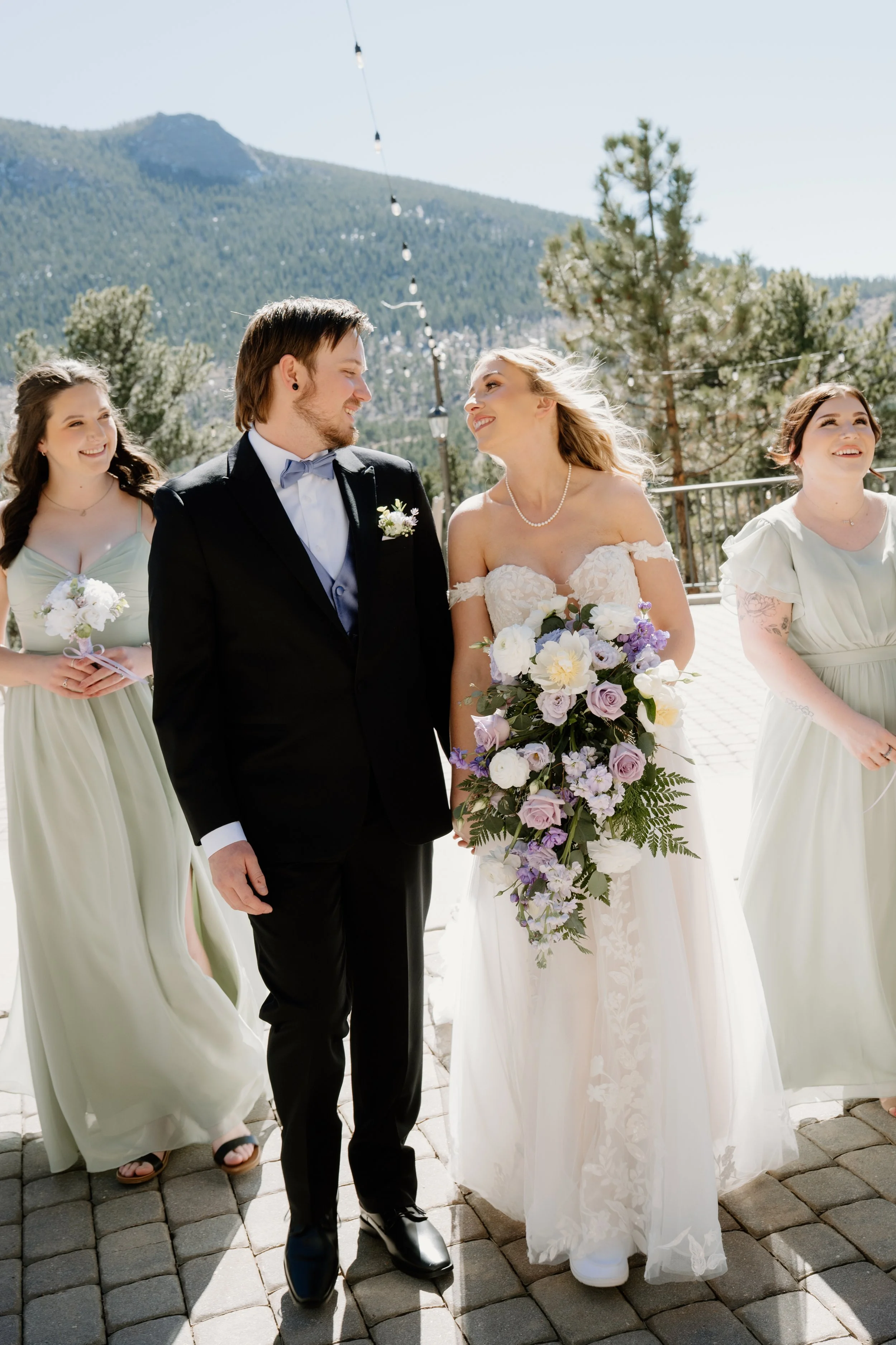 A couple holding hands looking at each other lovingly and their bridal party walking behind them, wedding colors are sage green and lilac