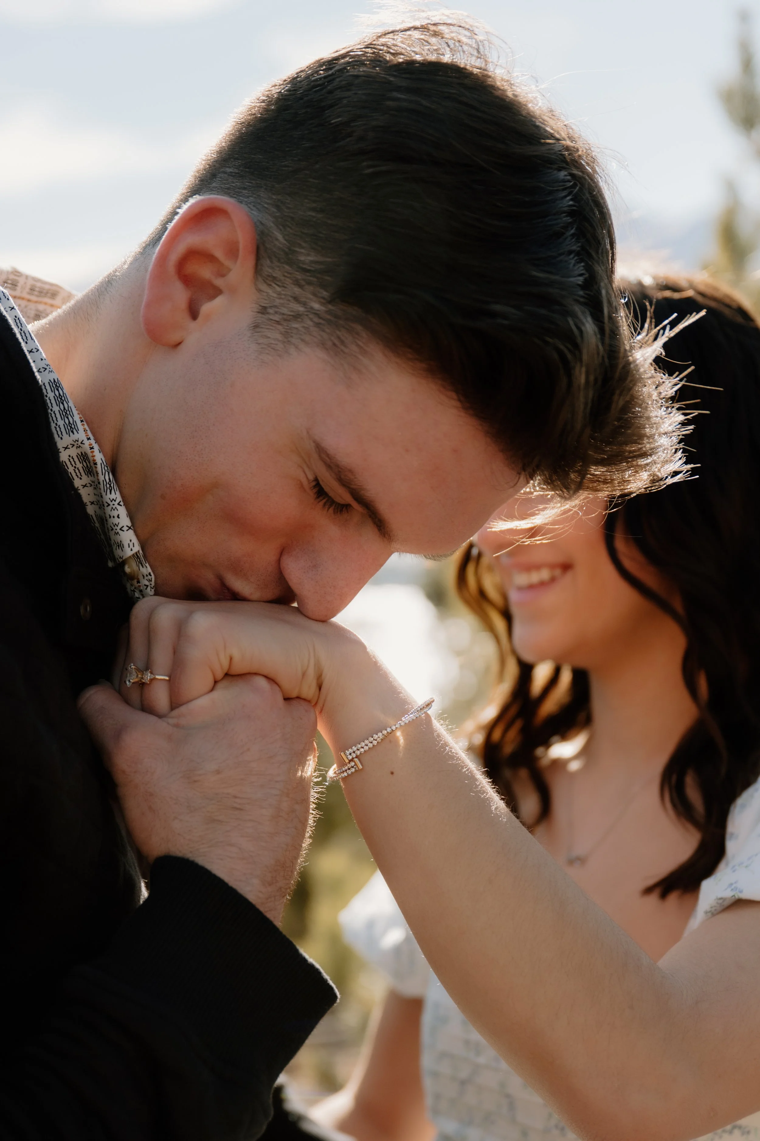 A couple celebrating their engagement at Dillon Lake in Elizabeth Colorado, man kissing fiancé's ring hand, laughing together