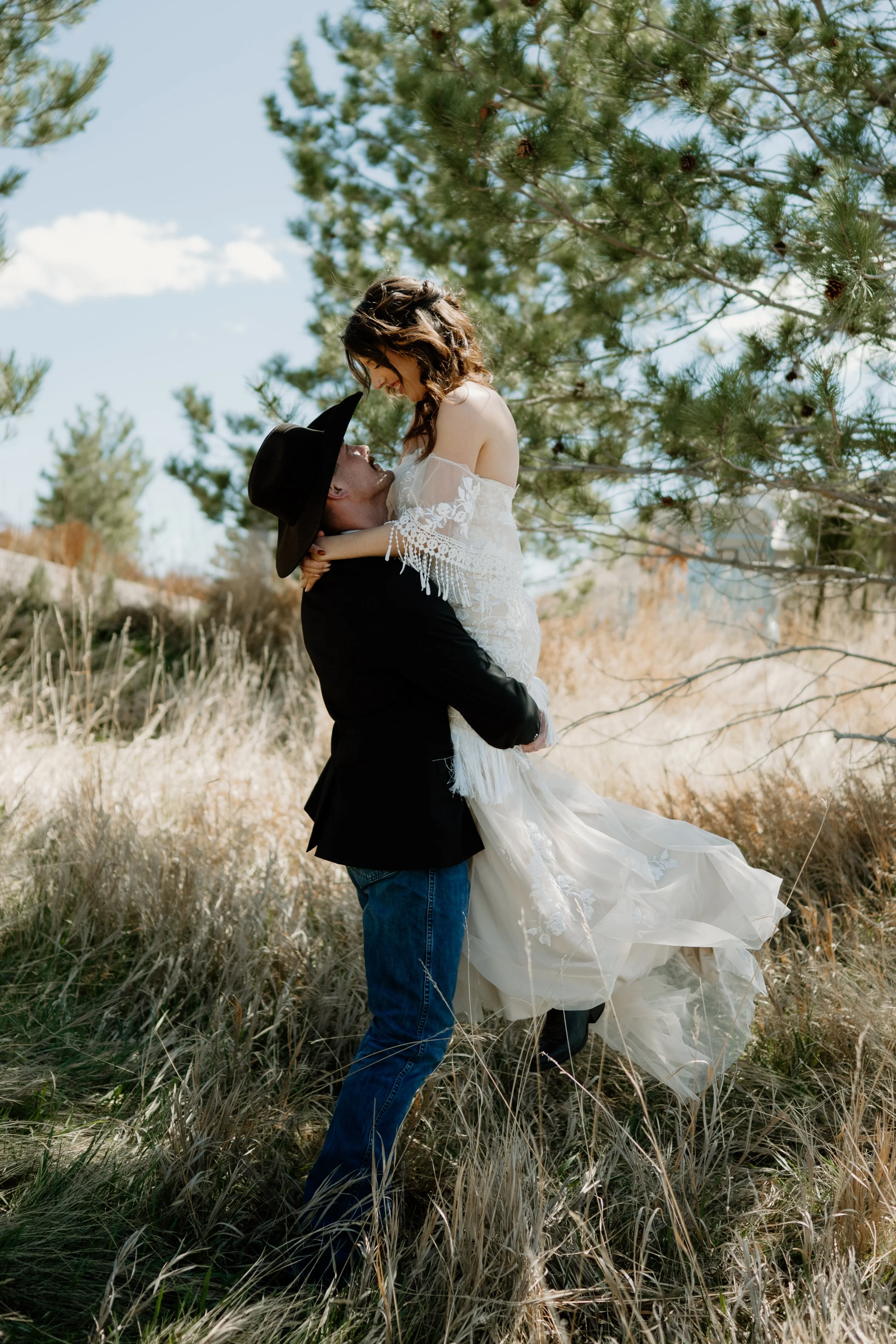 A western couple celebrating after their ceremony, groom holding bride up in the air with jeans, cowboy boots, and cowboy hat
