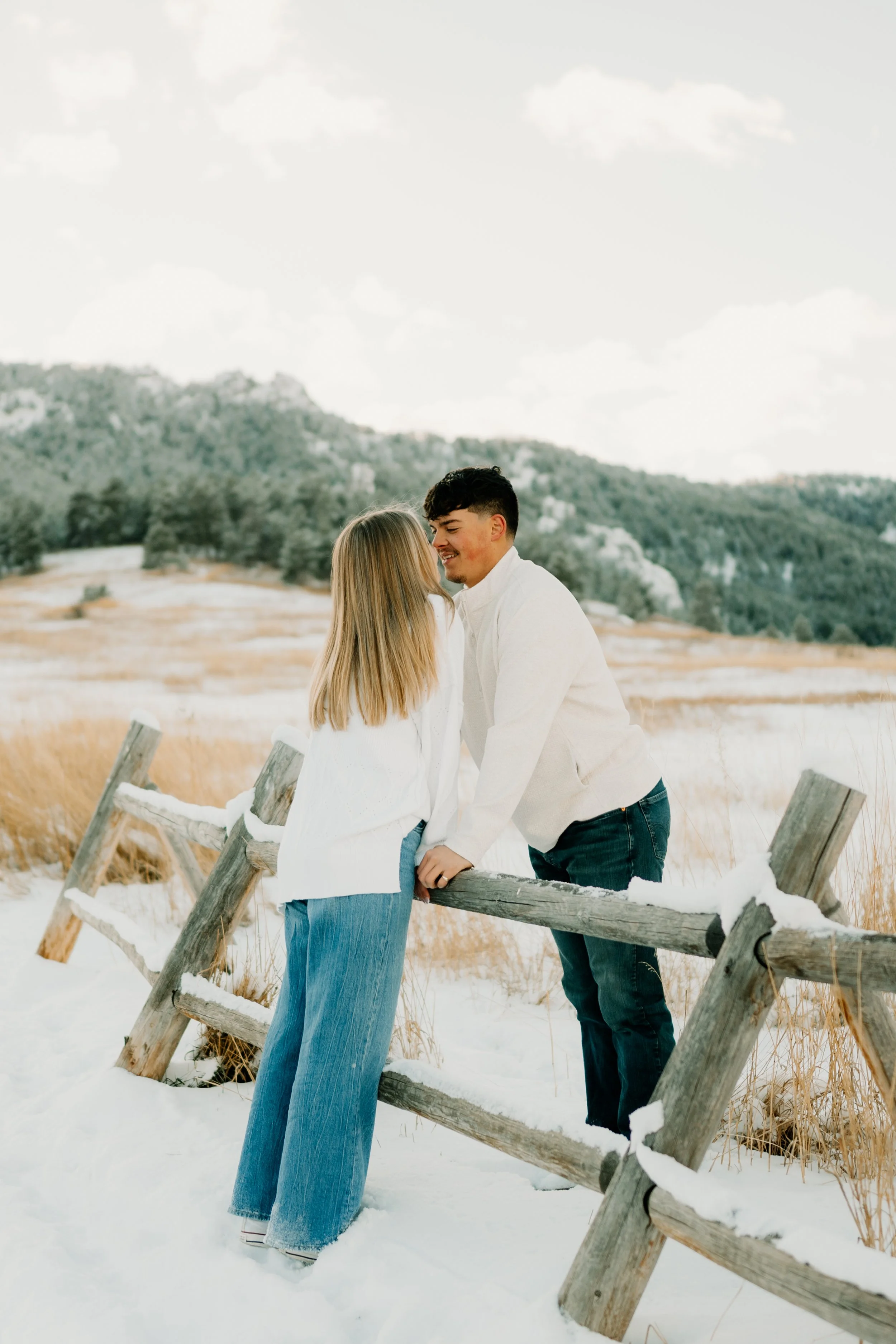 A couple celebrating their engagement in Boulder Colorado at Chautauqua Park leaning over a fence to kiss, both wearing jeans and a white top with snow on the ground