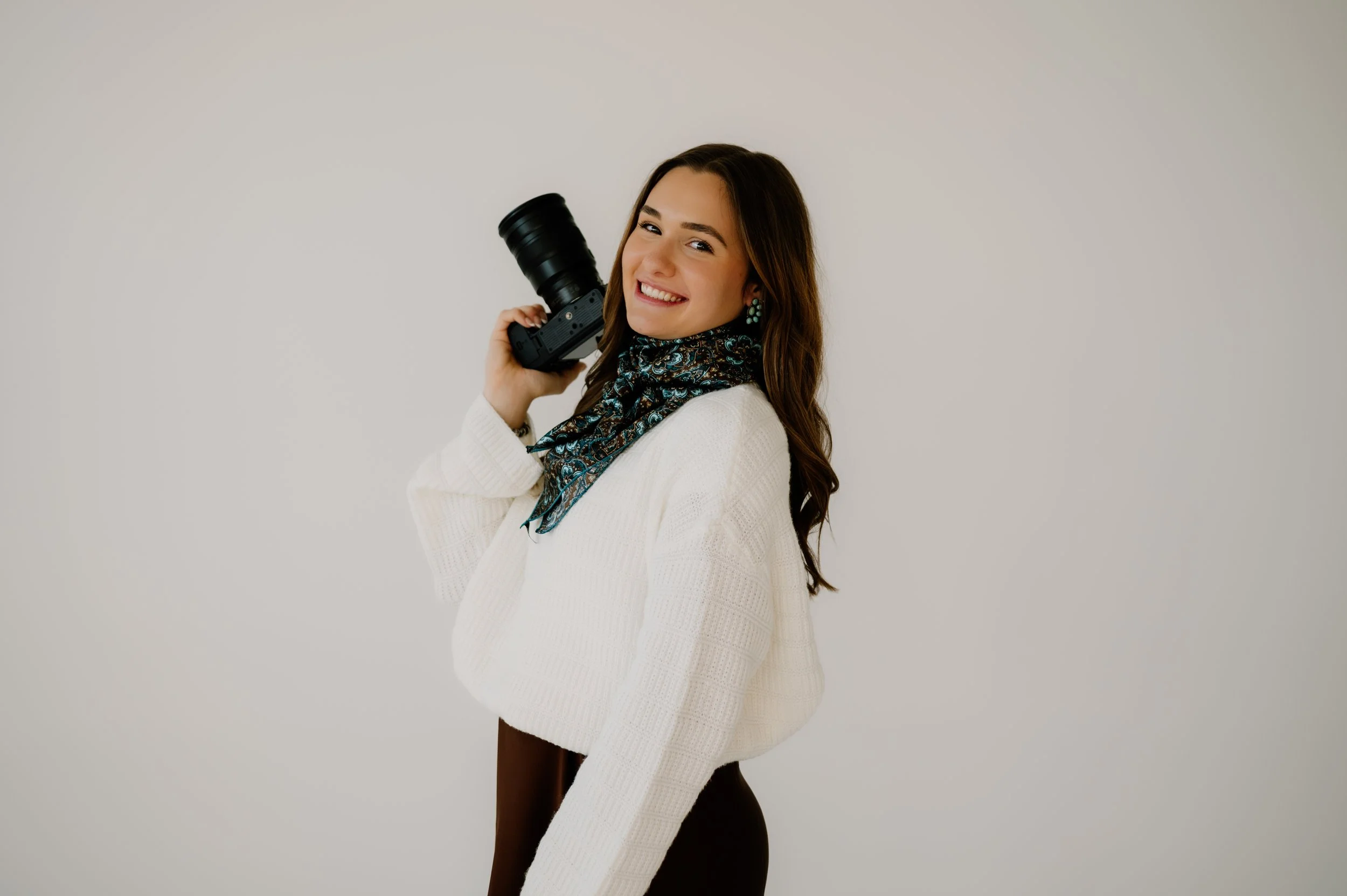 Western wedding and portrait photographer wearing a brown skirt and white sweater with a brown and blue wild rag and a turquoise half cluster earrings holding a camera