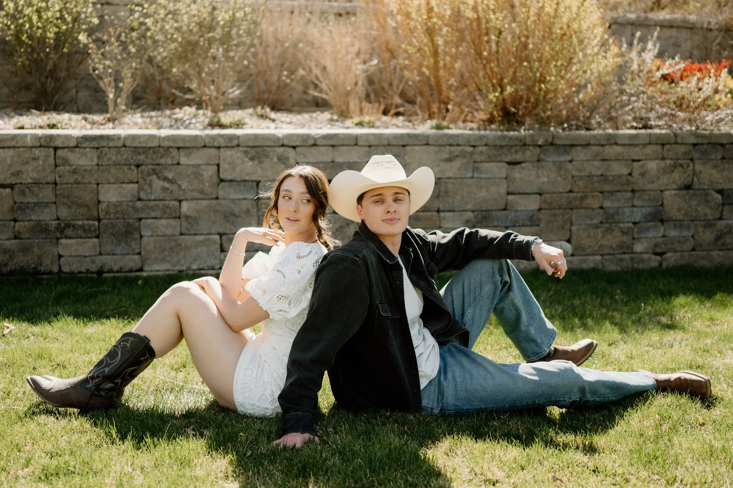 An engaged couple sitting back to back in the grass, girl looking over shoulder at fiancé, wearing cowboy boots and short white lace dress