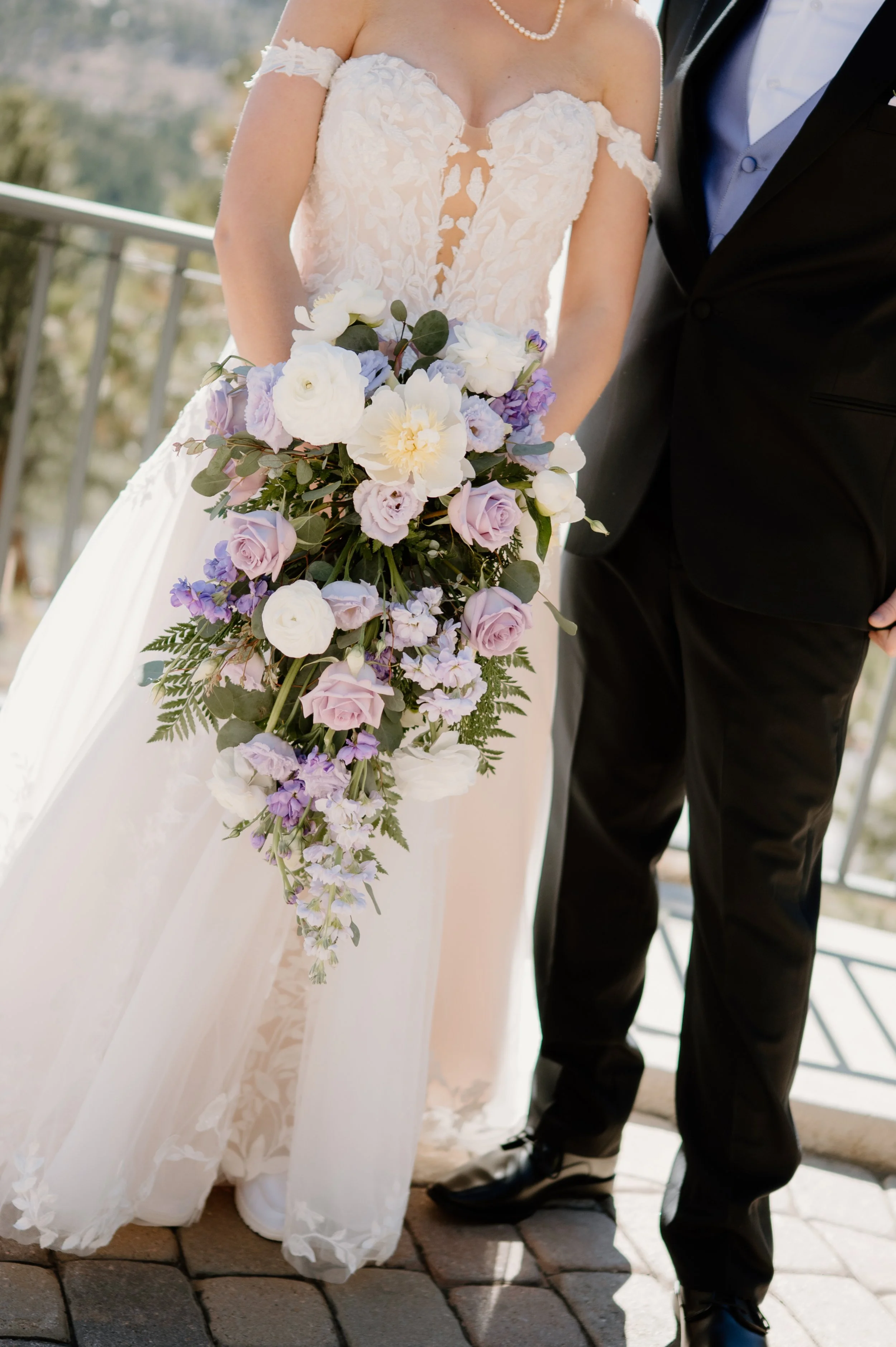 A just married couple holding wedding bouquet of lilac and white roses and florals, standing in front of the Colorado mountains in Estes Park Colorado at Della Terra 