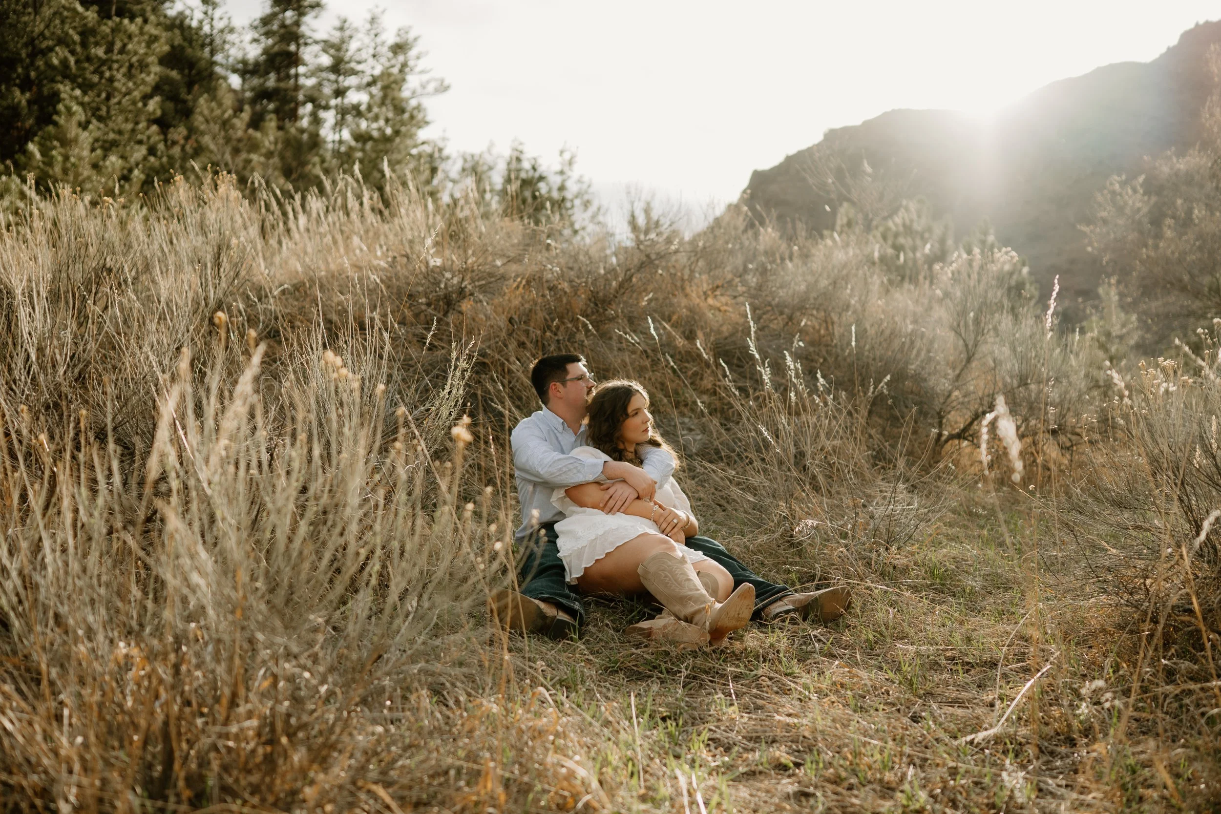 Engaged western couple sitting together in green grass in Loveland Colorado, girl is wearing a white dress and tall cowboy boots