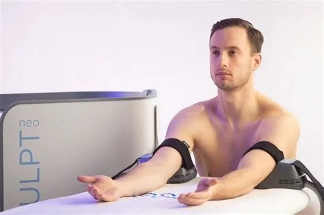 A man undergoing a medical test with a device wrapped around his arm, sitting at a medical examination table.