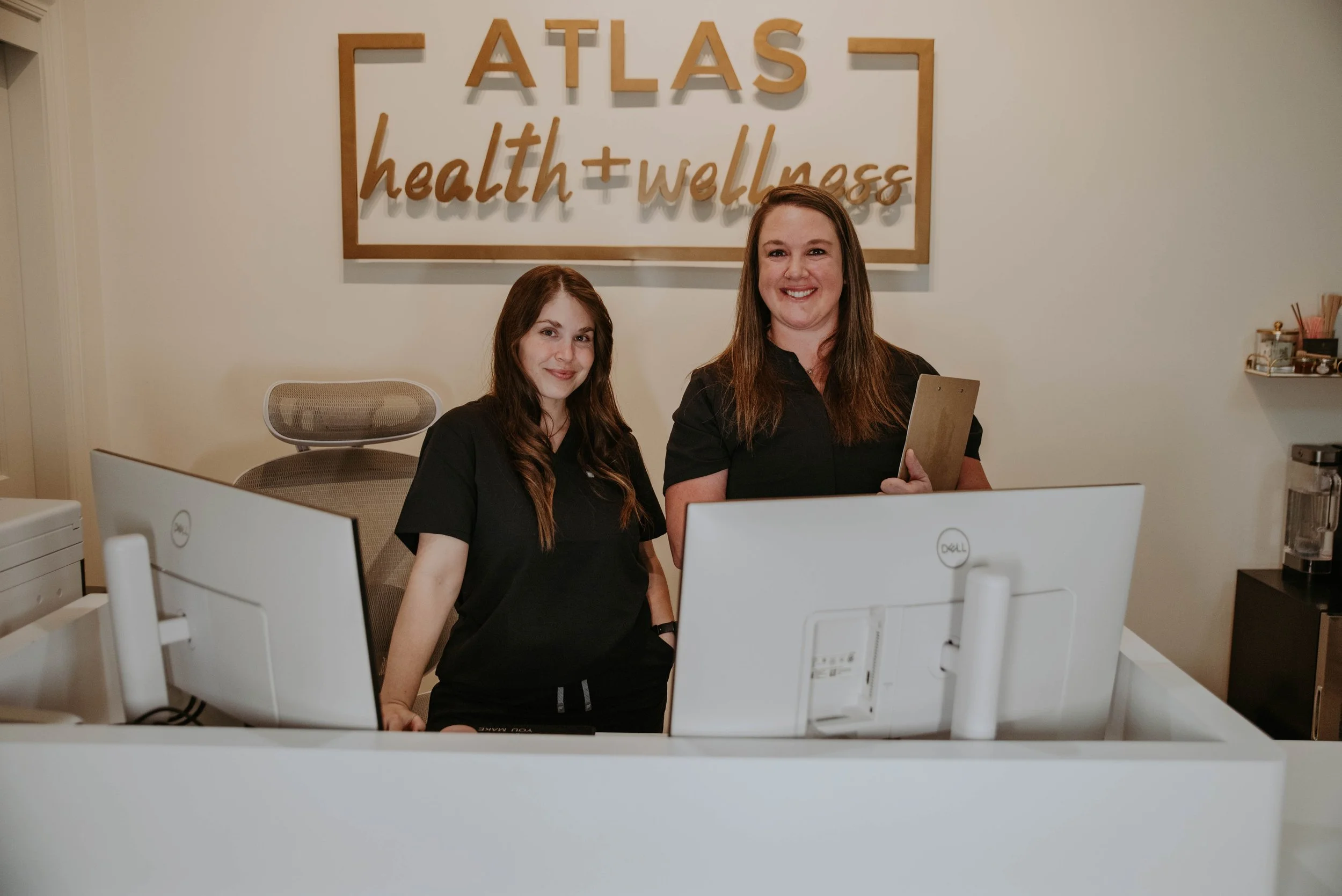 Two women smiling at a desk with desktop computers in an office. A sign on the wall reads 'ATLAS health + wellness.'