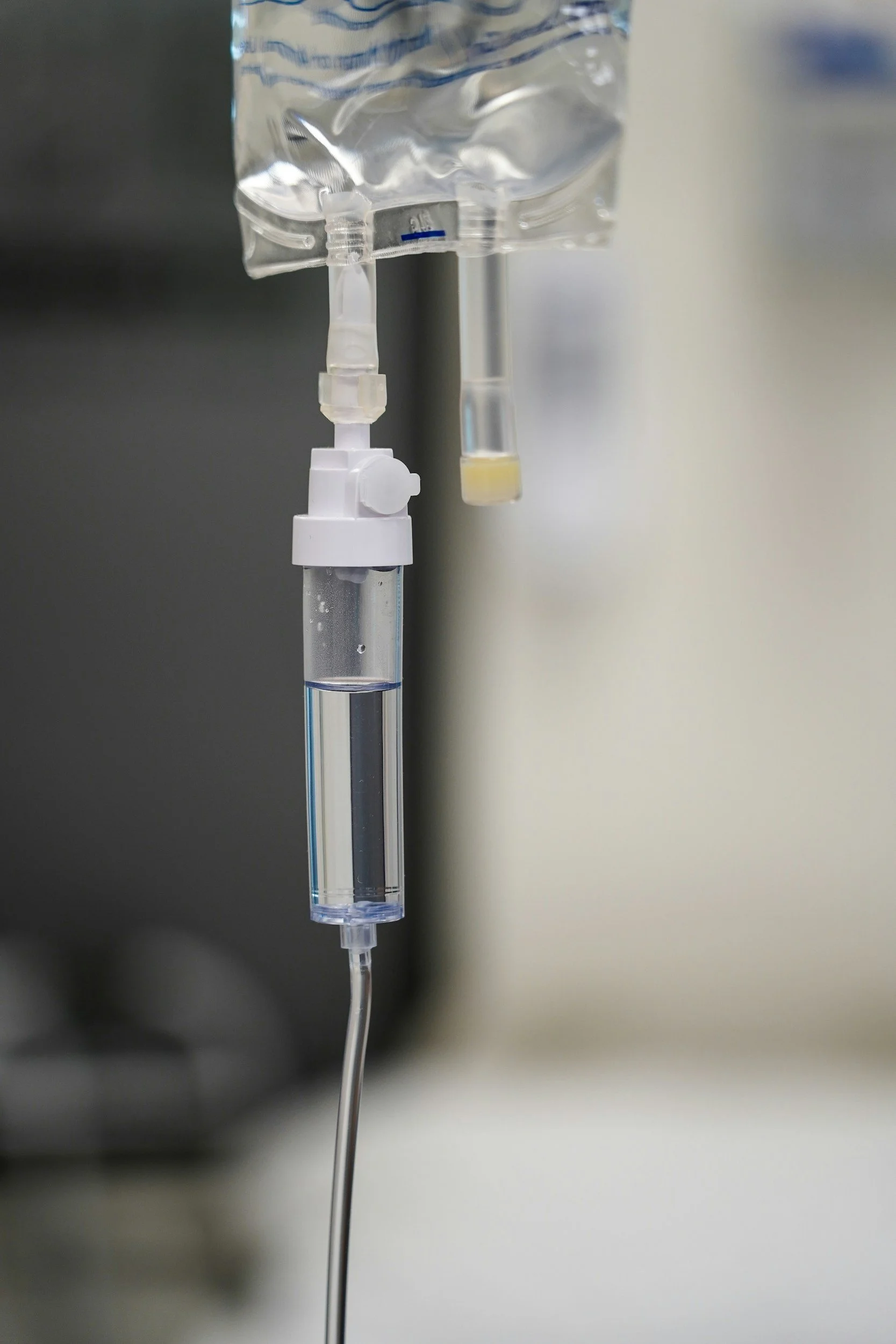 A woman wearing a black shirt and pink gloves is preparing an IV drip. She is adjusting the IV bag hanging on a stand in a room with white panel walls.